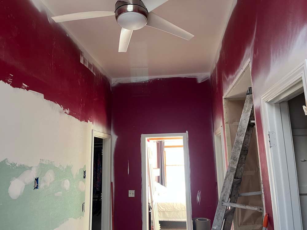 Partially painted hallway with magenta walls, white ceiling fan, and ladder near a doorway