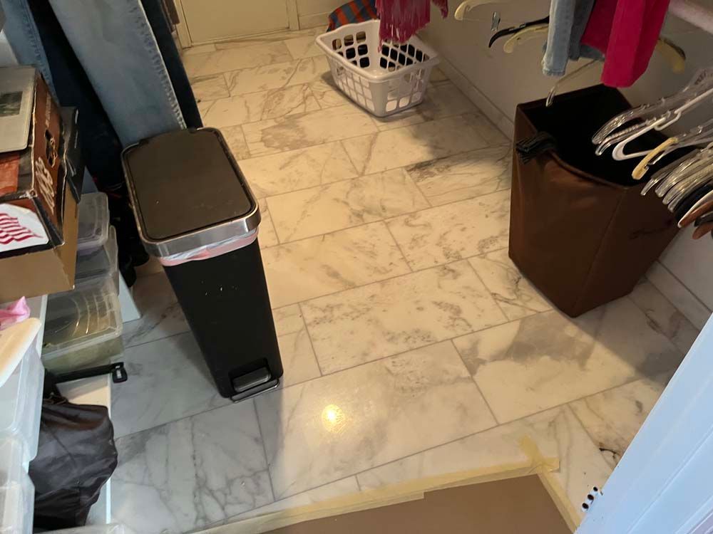 Small closet with marble tile floor, laundry basket, trash can, and hanging clothes.
