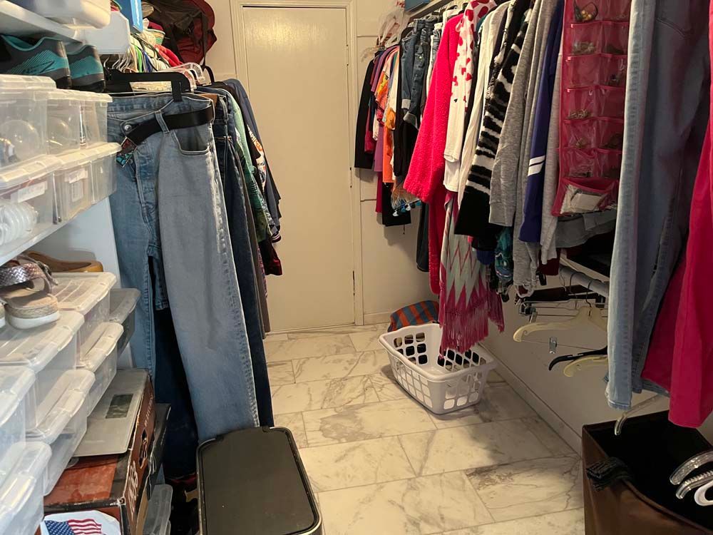 Narrow walk-in closet with hanging clothes, storage bins, shelves, and a laundry basket on the floor