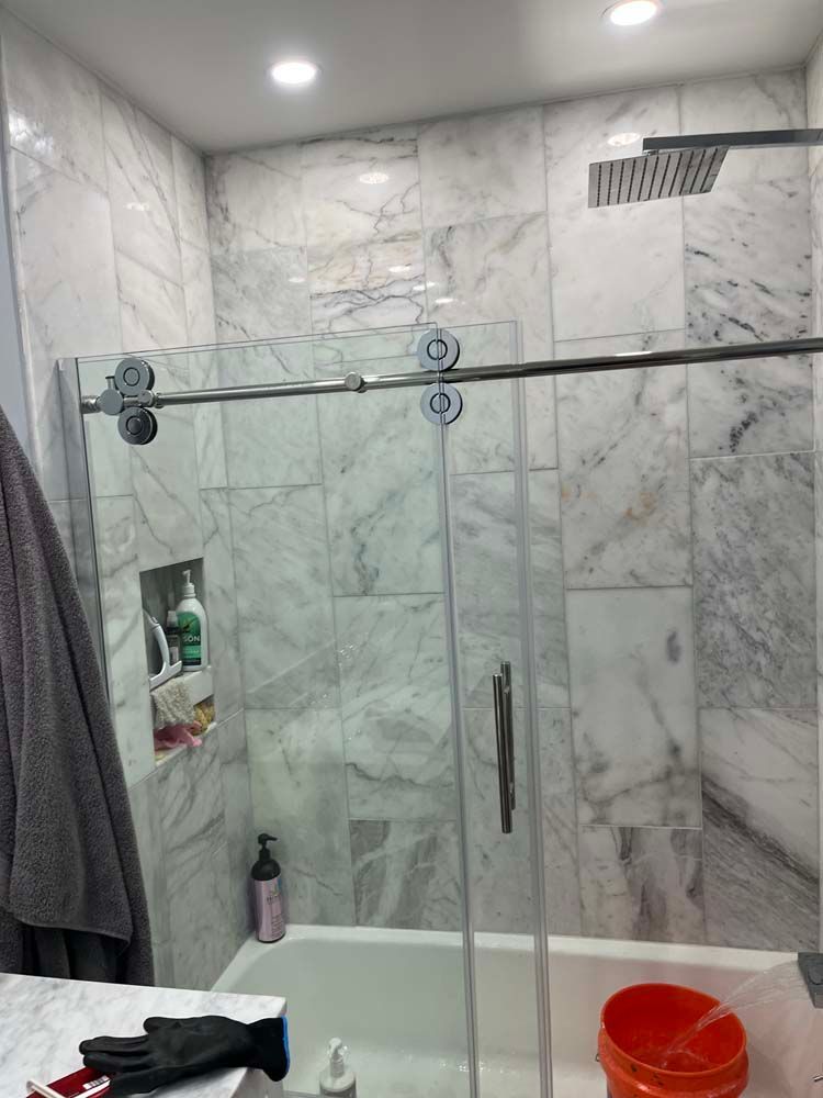 Modern bathroom with glass shower enclosure, gray marble walls, towel, shelf niche, and orange bucket.