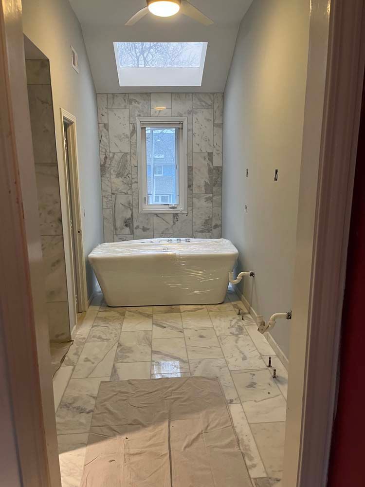 Narrow bathroom hallway with marble tile, a freestanding tub, skylight, and a window at the far end.