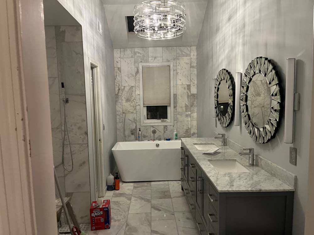 Modern bathroom with marble tile, double vanity, round mirrors, and a freestanding tub near a window.