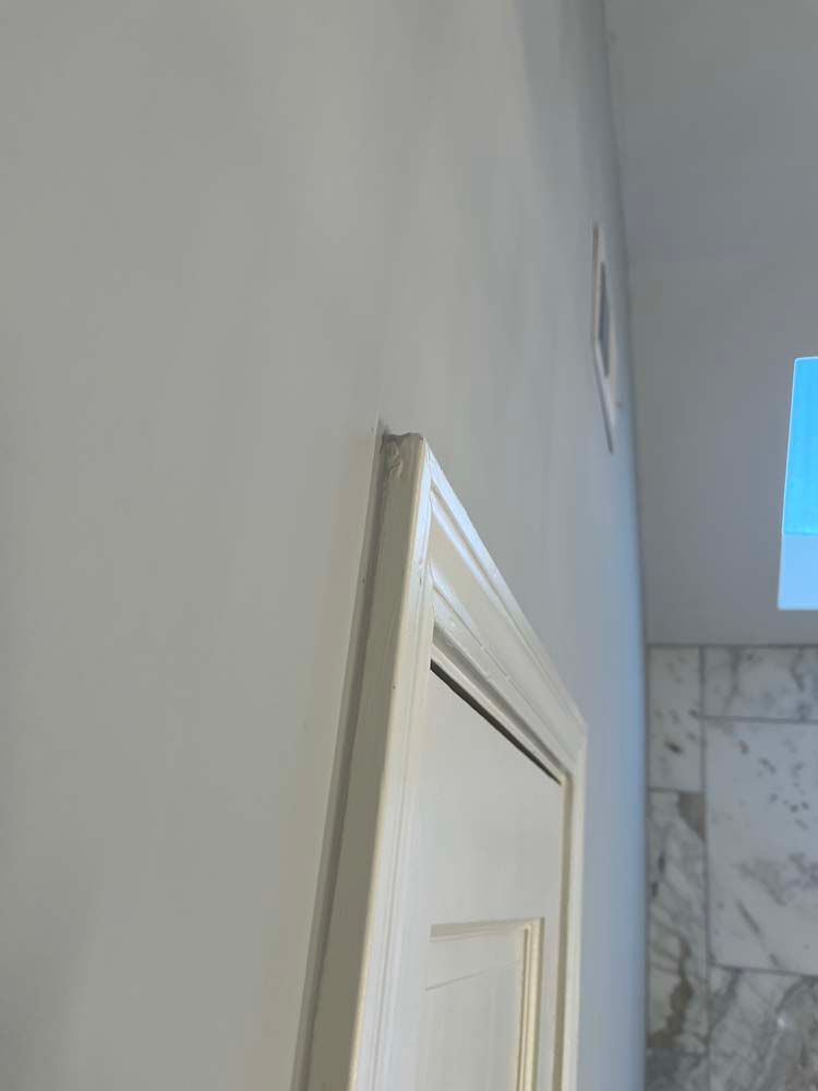 White interior door with trim beside a gray wall and marble-tiled shower area near a small window.