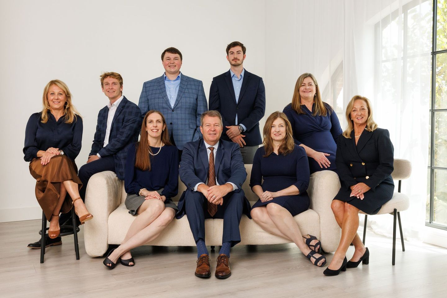 Seven people pose in a bright living room, seated and standing on a white sofa with large windows behind them.
