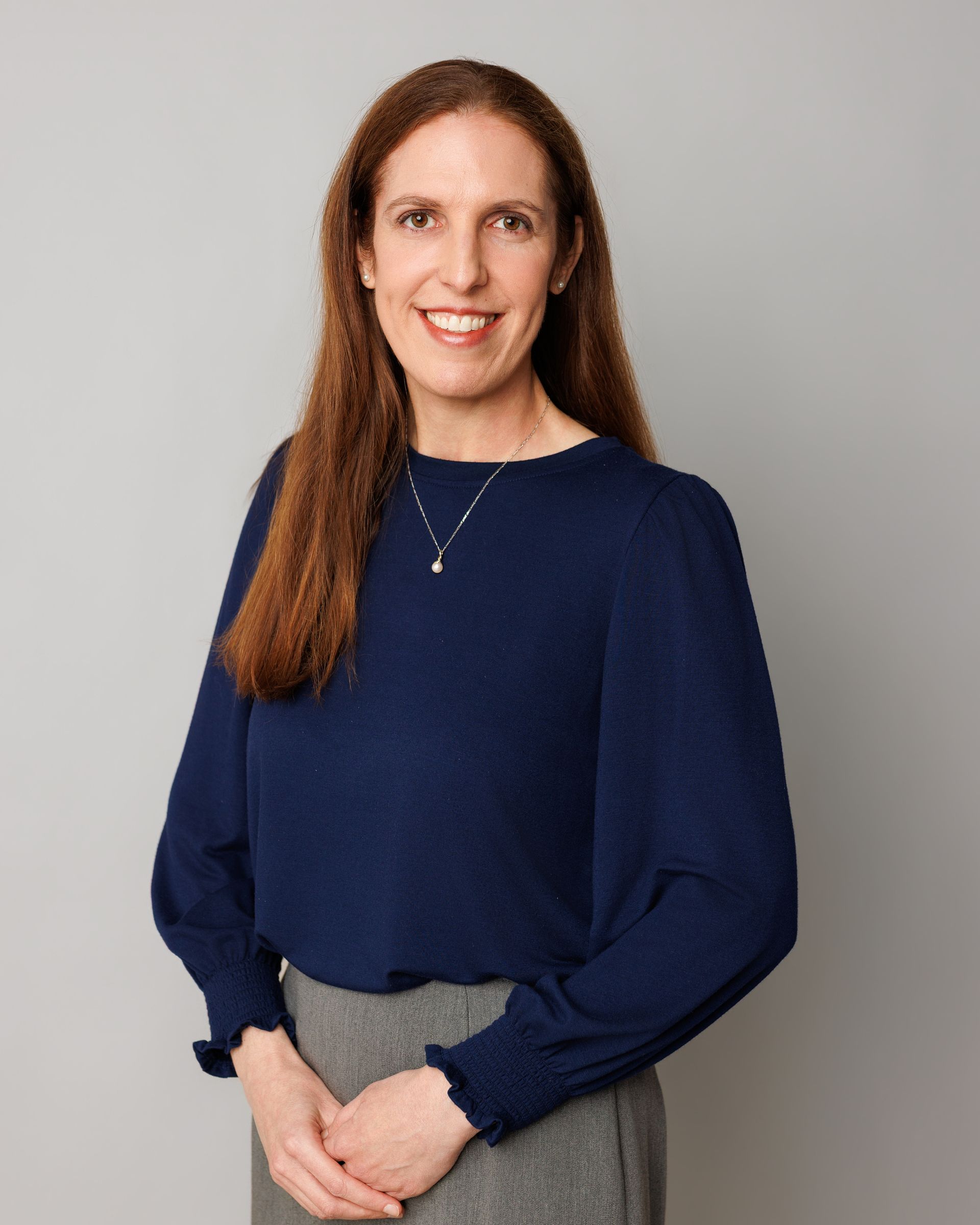 Woman standing against a gray background, wearing a navy blouse and smiling at the camera.