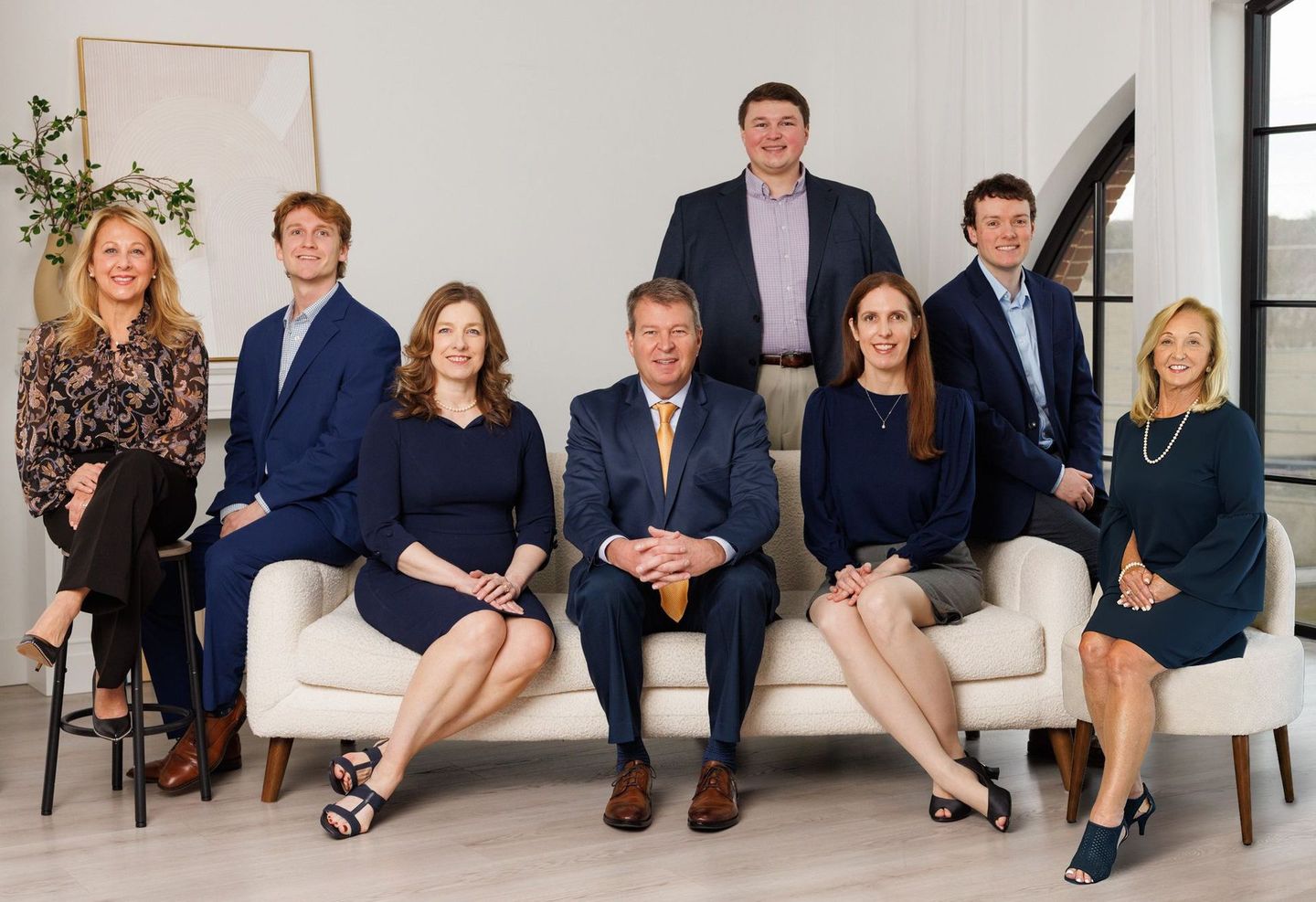 Seven people pose in a bright living room, seated and standing on a white sofa with large windows behind them.