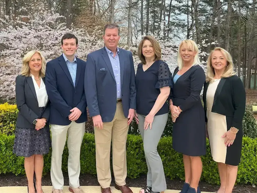 Six colleagues posing outdoors in business attire in front of trees and landscaping.