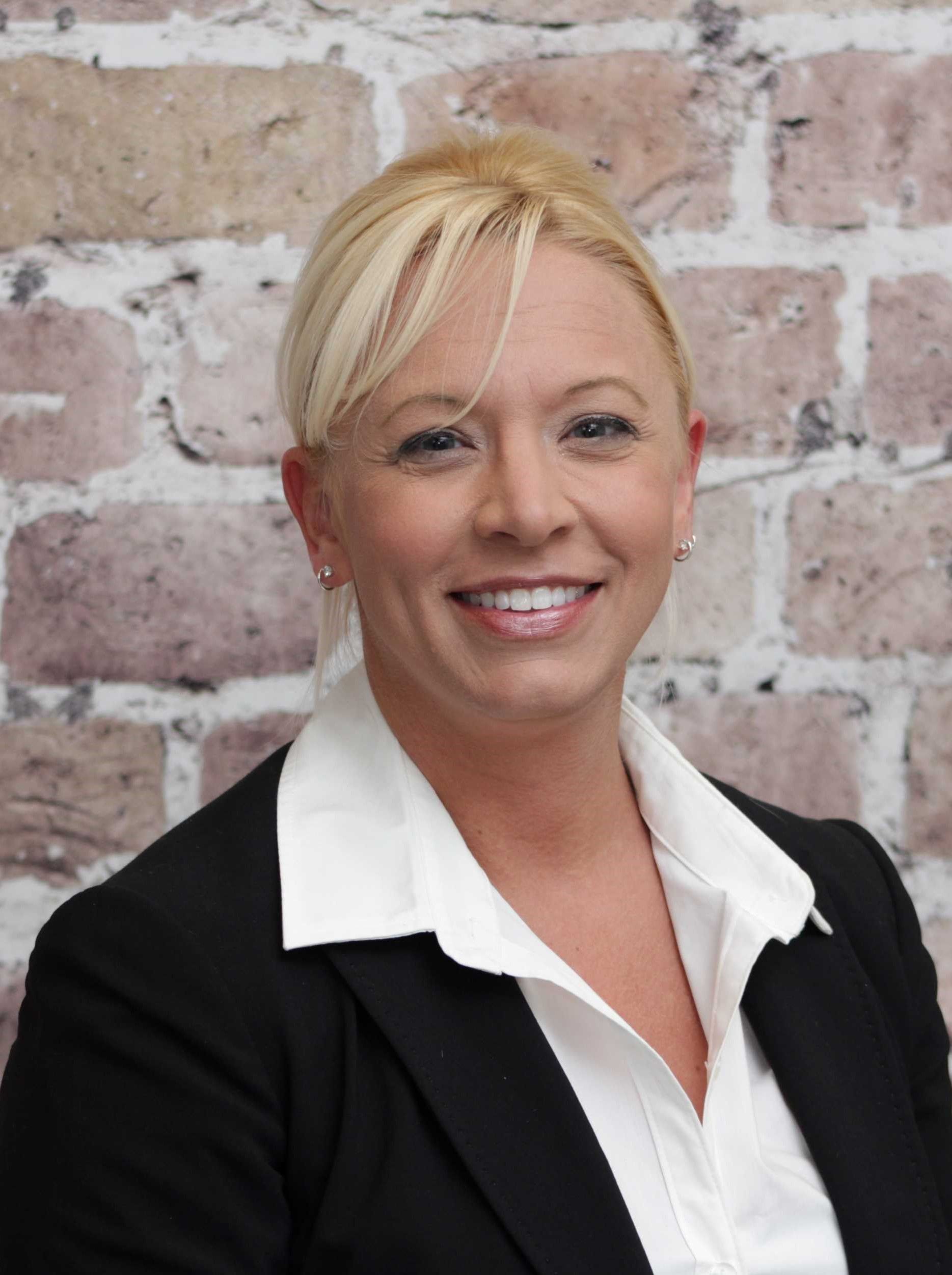 Smiling woman in a black blazer and white collared shirt against a brick wall