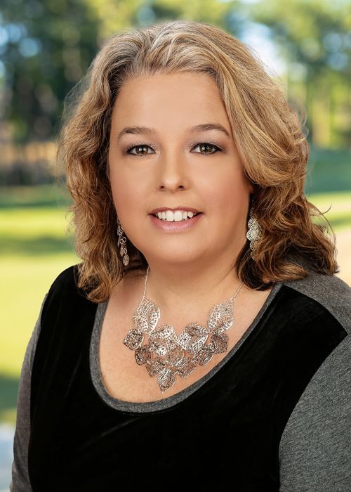Portrait of a smiling woman wearing a black top and silver necklace, with a blurred outdoor background