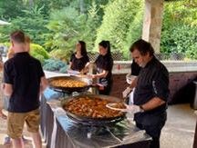 People serving food at large trays during an outdoor buffet under a pavilion
