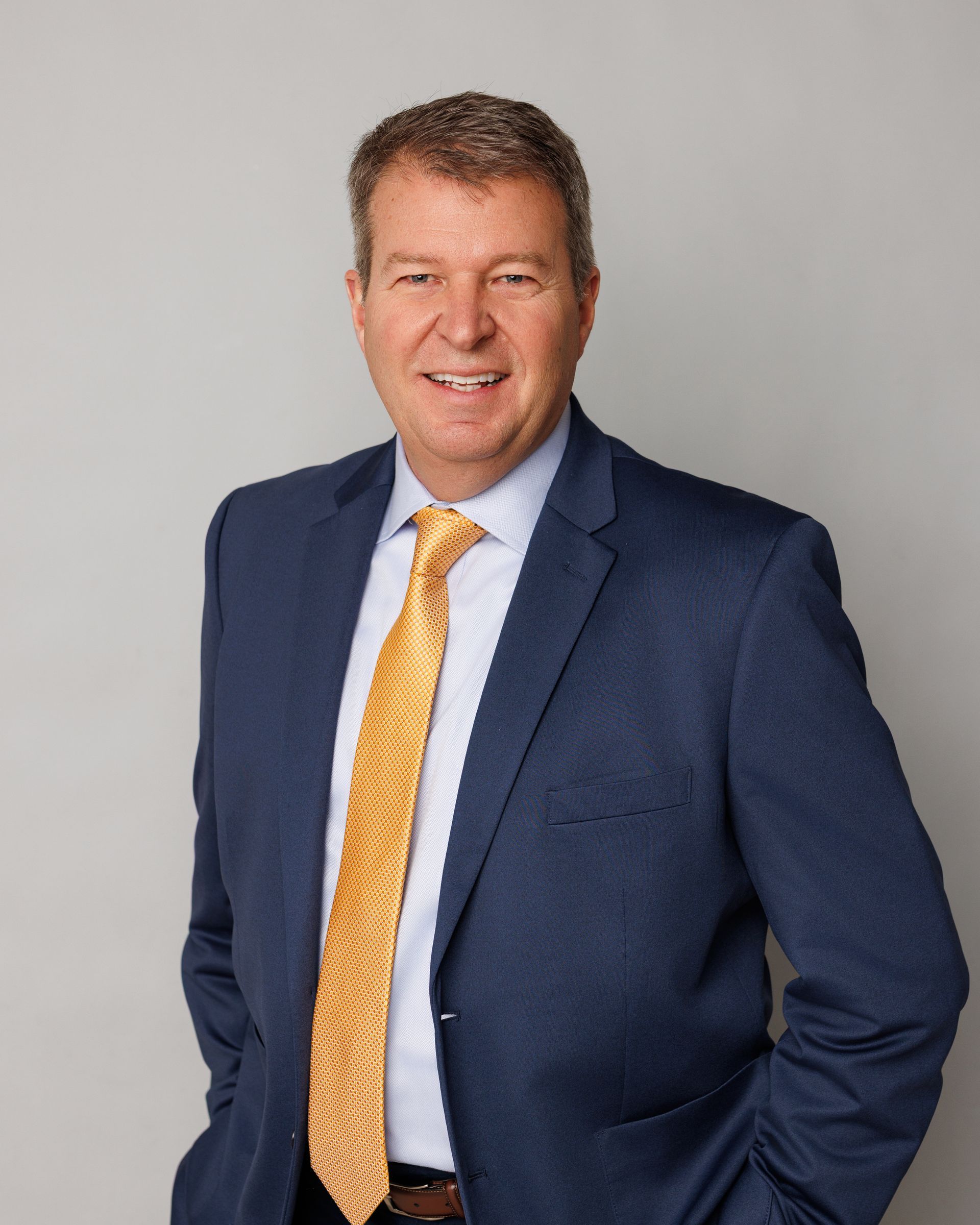 Man in a navy suit and yellow tie, smiling against a plain gray background