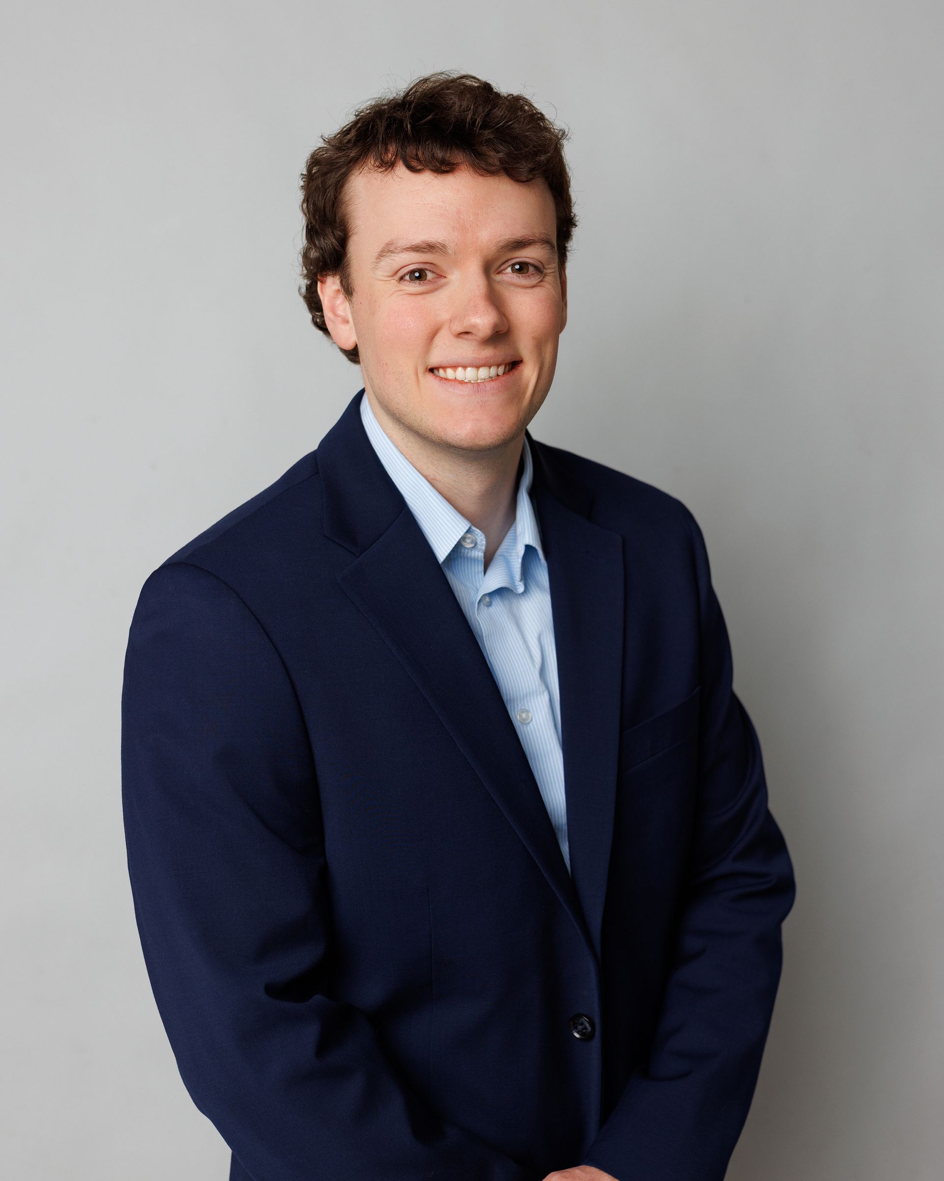 Man in a navy blazer and light blue shirt smiling against a gray studio background