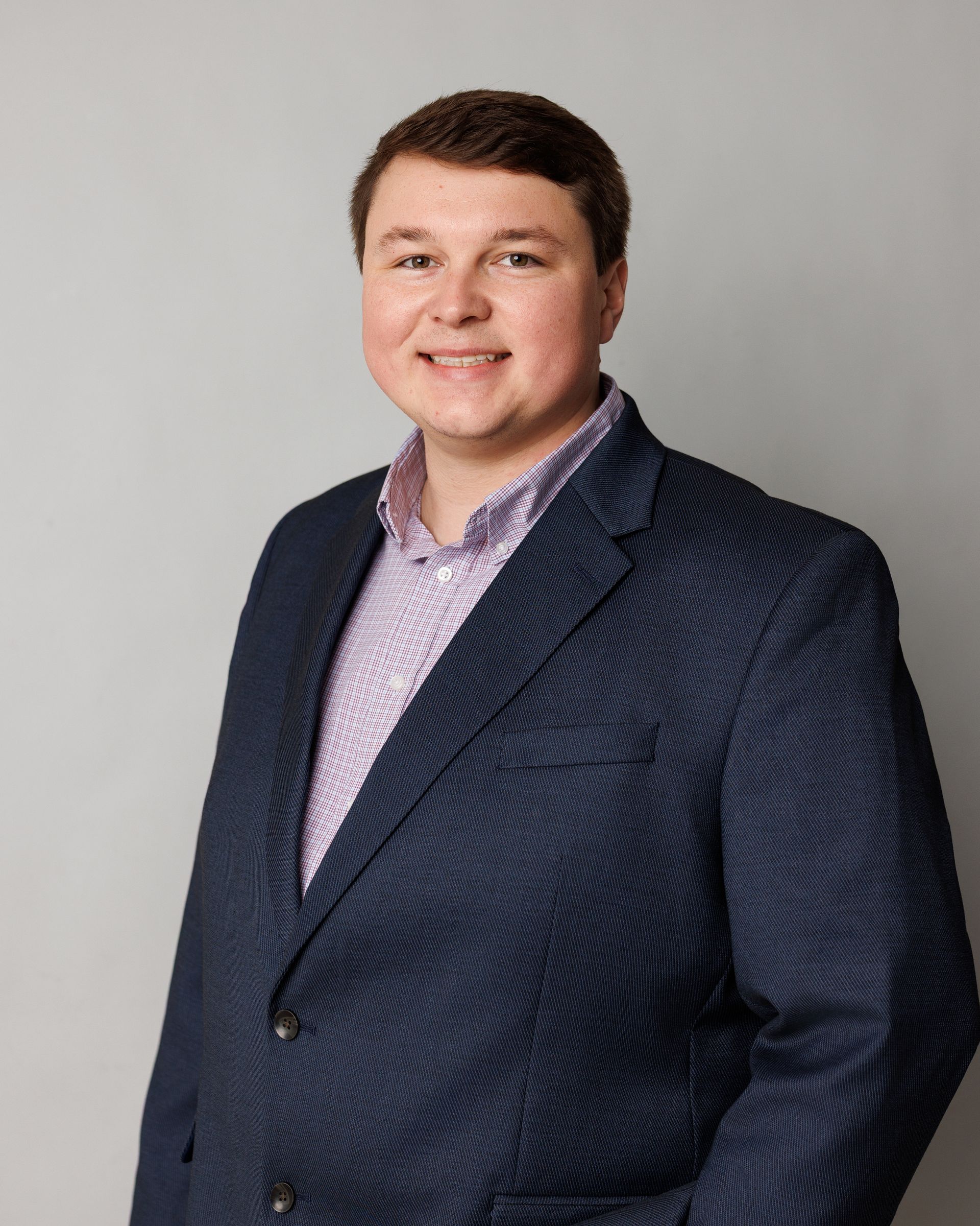 Smiling person in a navy blazer and purple checkered shirt against a gray background