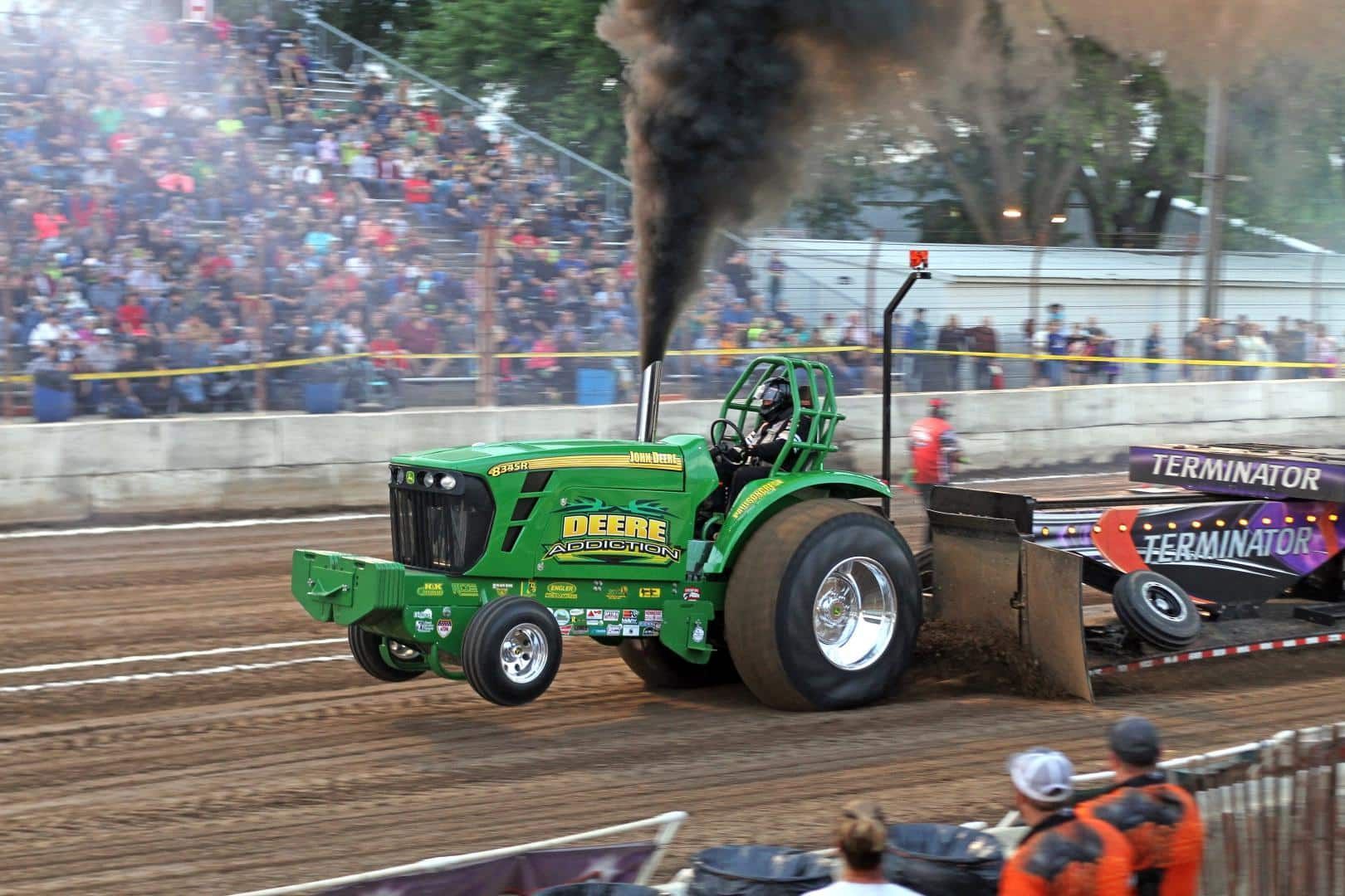Green tractor pull truck racing on dirt track, black smoke billowing, with crowd in stands behind it