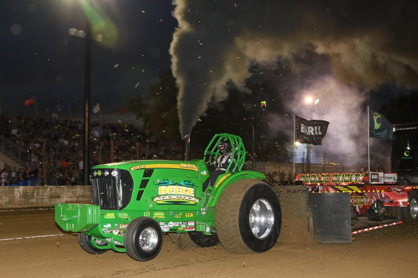 Green tractor pulling a sled on a dirt track at night, with smoke rising and a crowd in the stands.