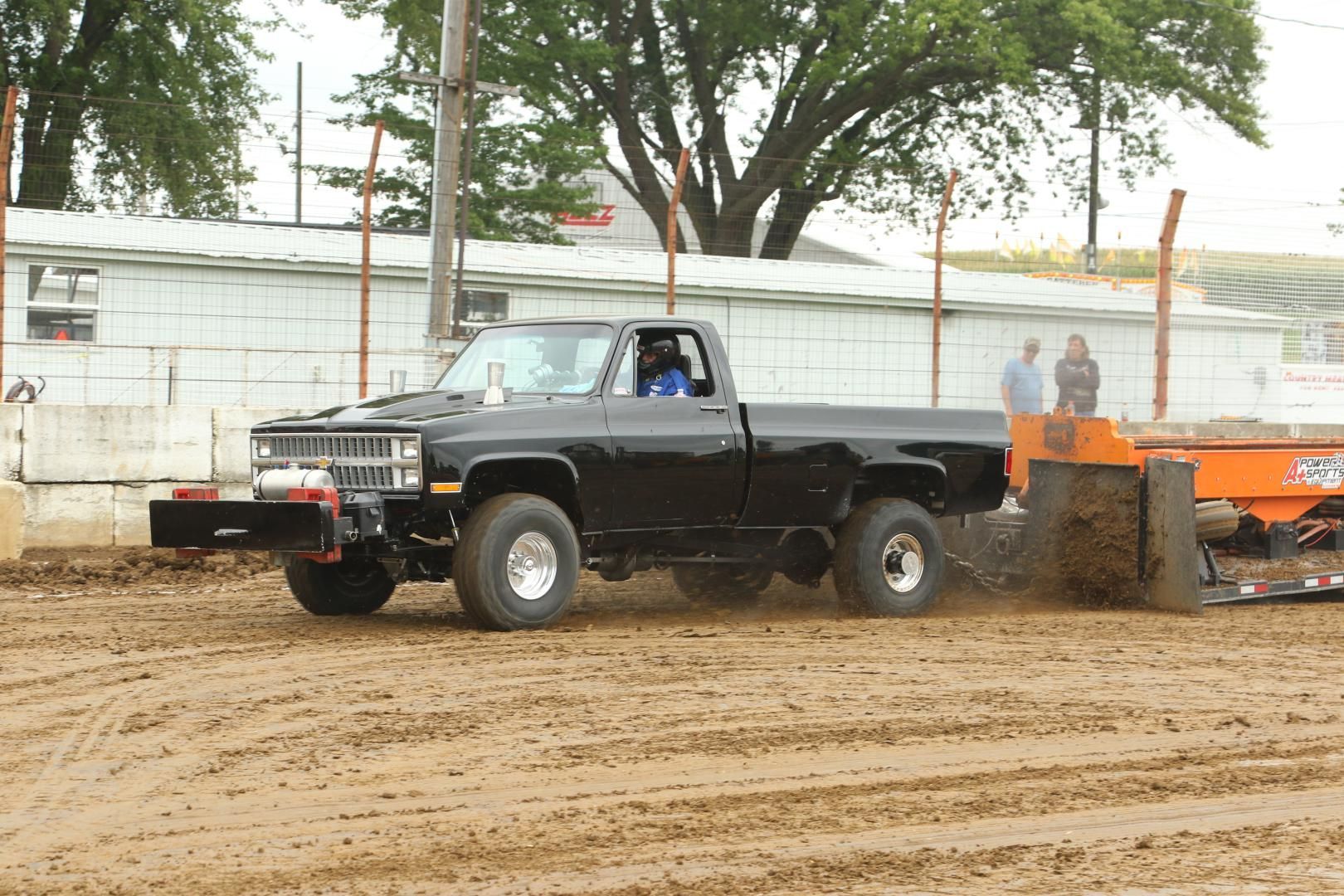 Black pickup truck pulling orange sled on muddy dirt track at a tractor pull event