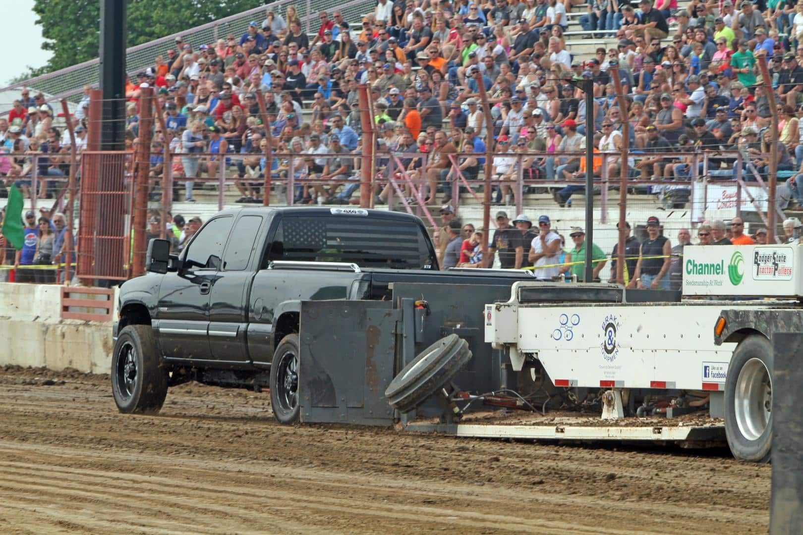Black pickup truck towing a white sled on a dirt track, with a packed grandstand in the background