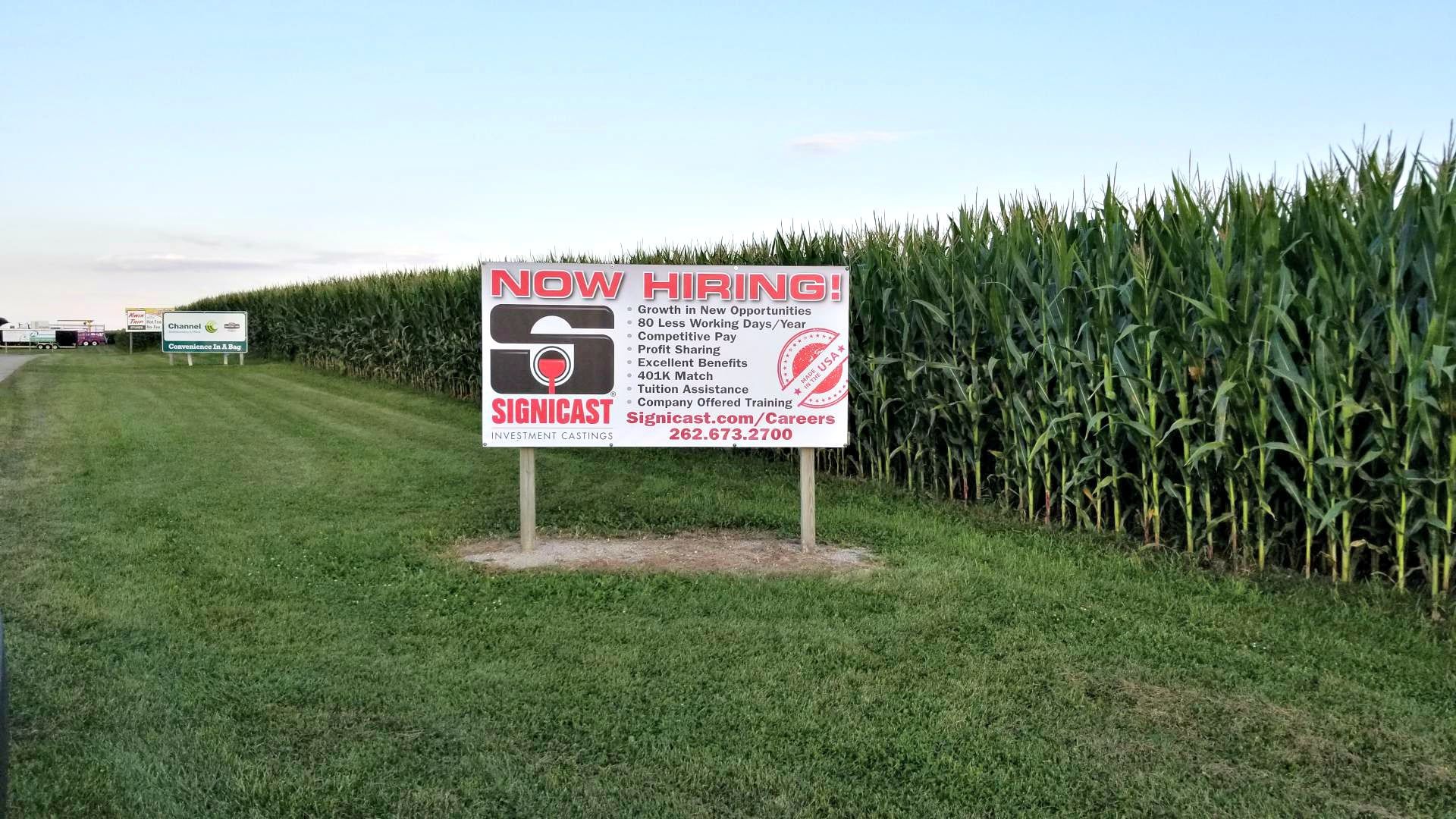 “Now Hiring” sign in a grassy field beside tall corn, with a road and sky in the background