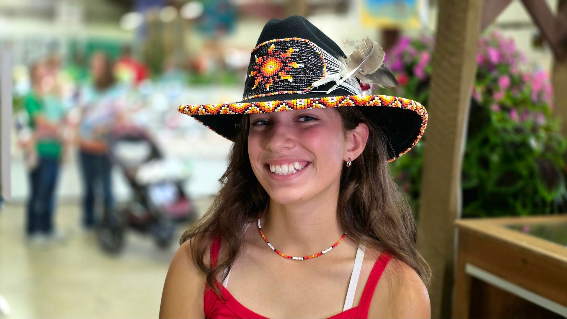 Smiling woman in a black floral hat and red top outdoors at a lively market.