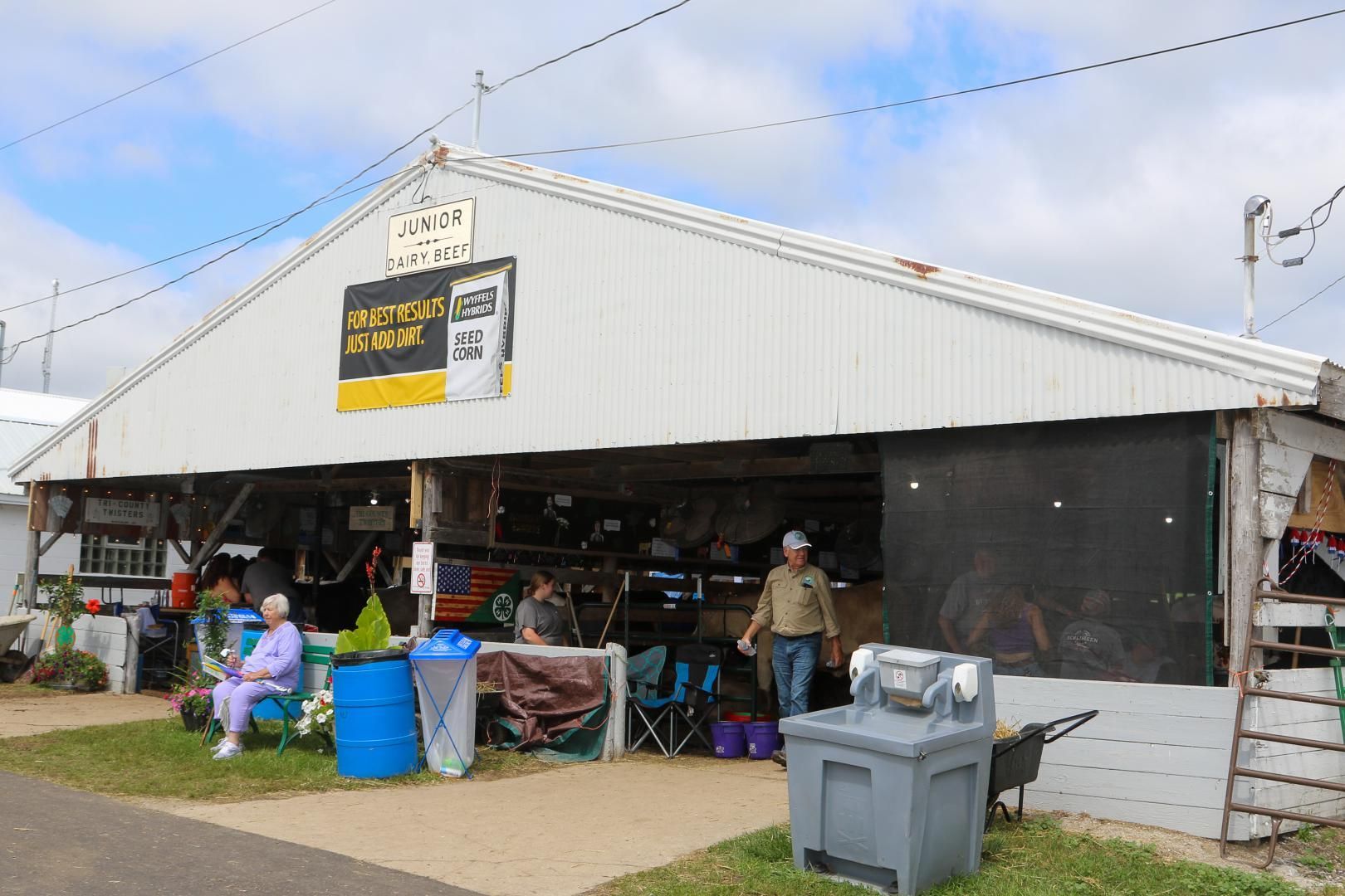 White market stall with people seated outside and goods under a canopy on a cloudy day