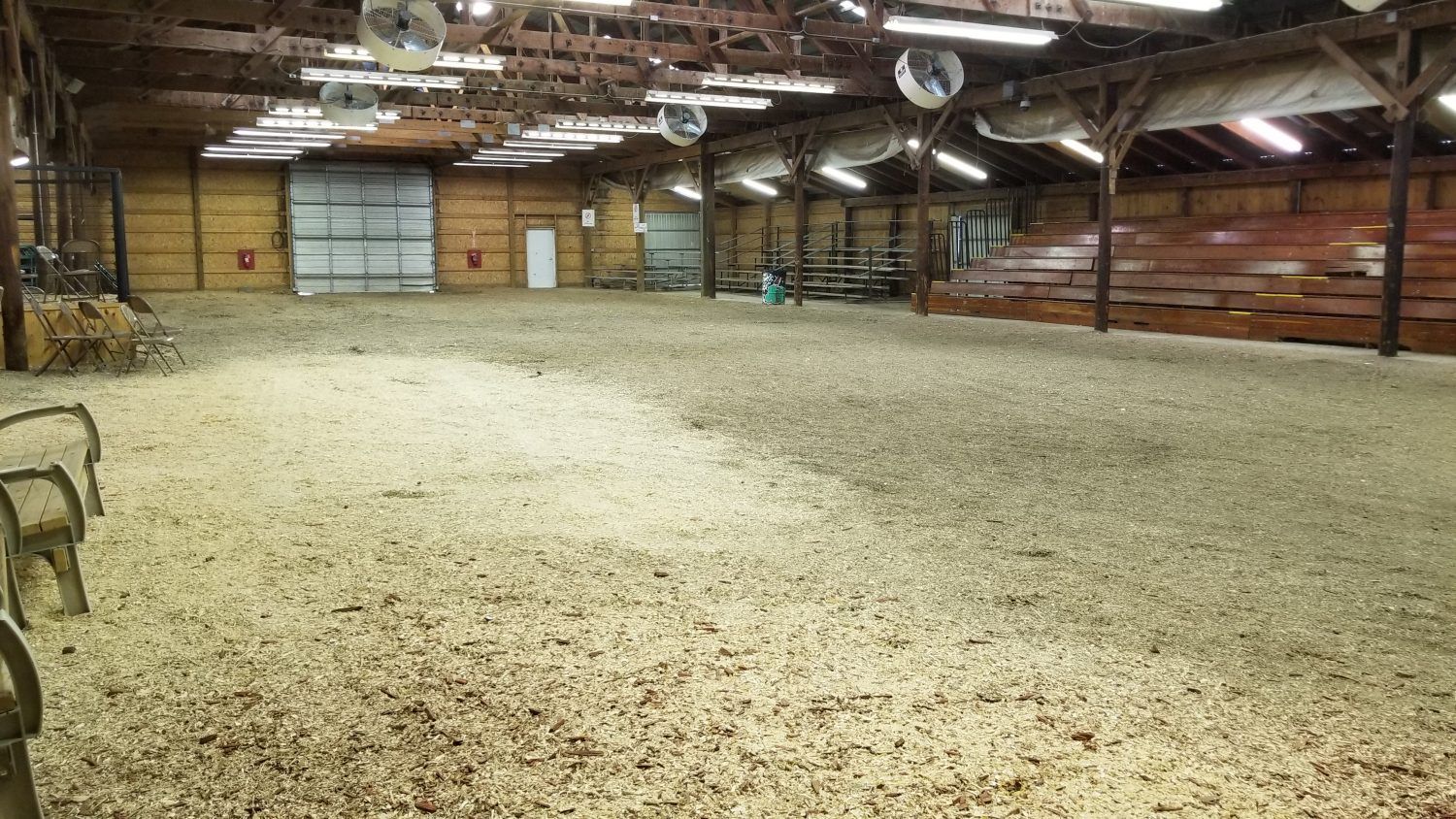 Empty indoor arena with dirt floor, wooden walls, overhead lights, and a large garage door in the back