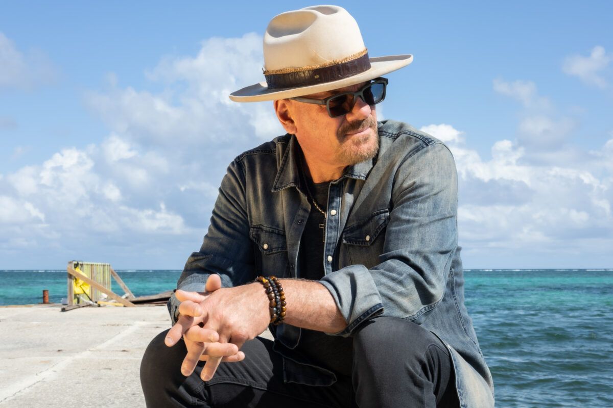 Man in a straw hat and denim shirt sitting on a beach with ocean and sky behind him