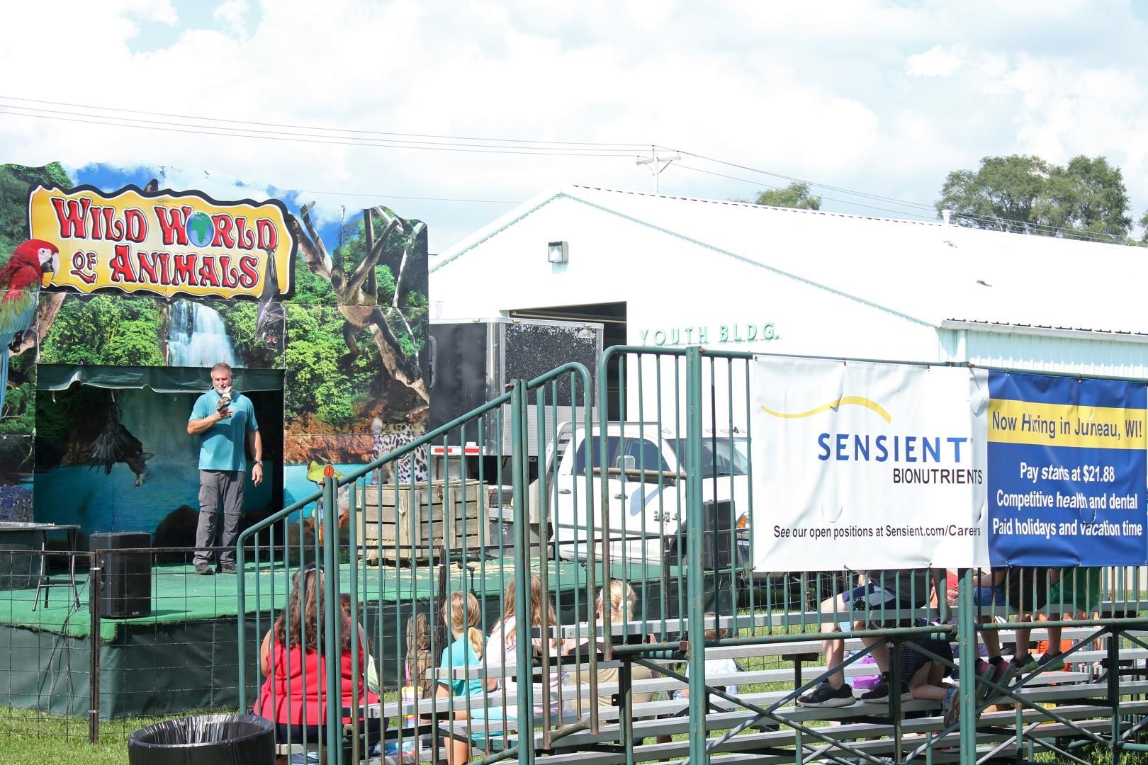 Outdoor stage with a person speaking beside a colorful “Wild Wild Wet” sign and a white “SENSIENT” tent.