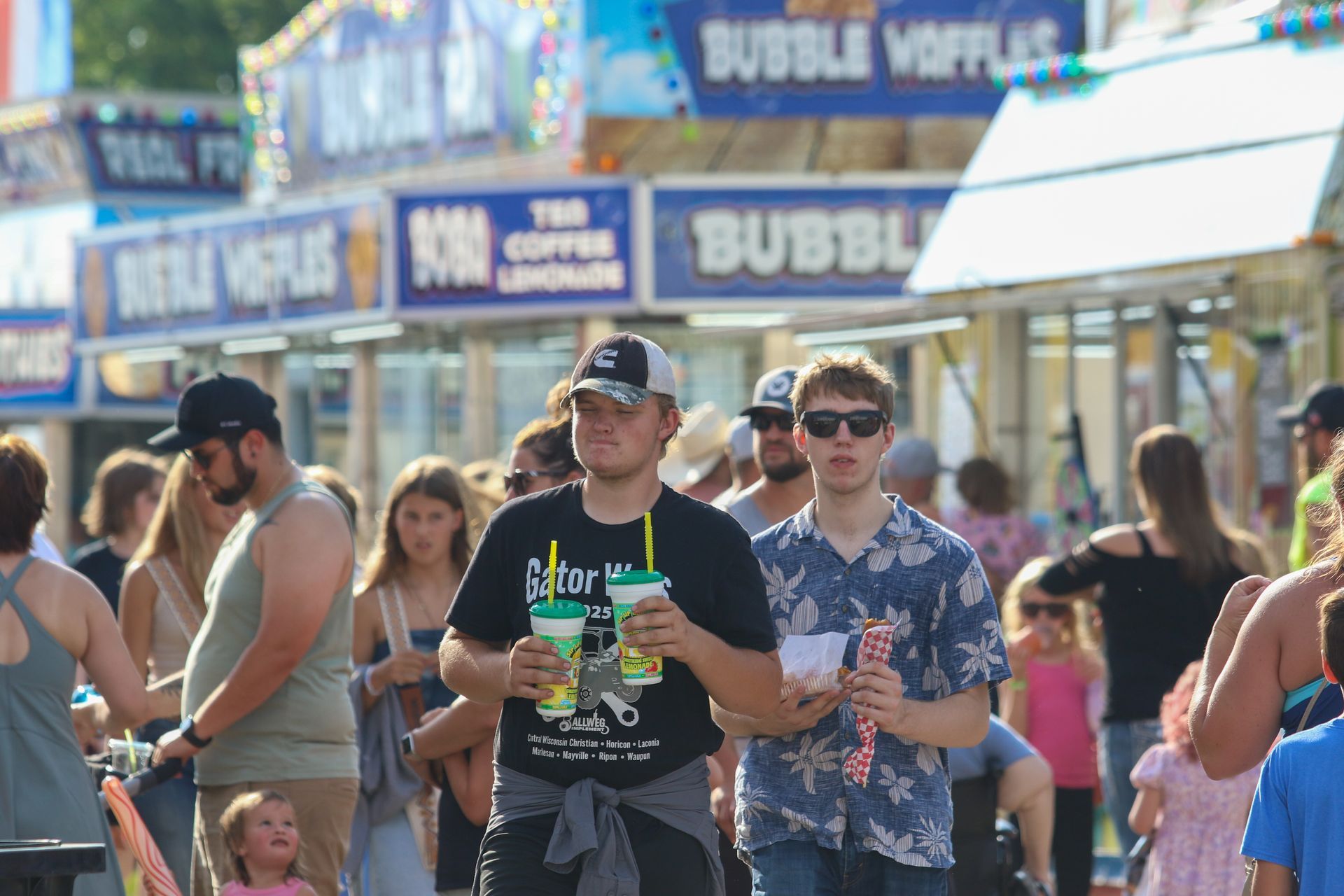 People walking past a bubble waffle stand at an outdoor fair, holding drinks in bright sunlight