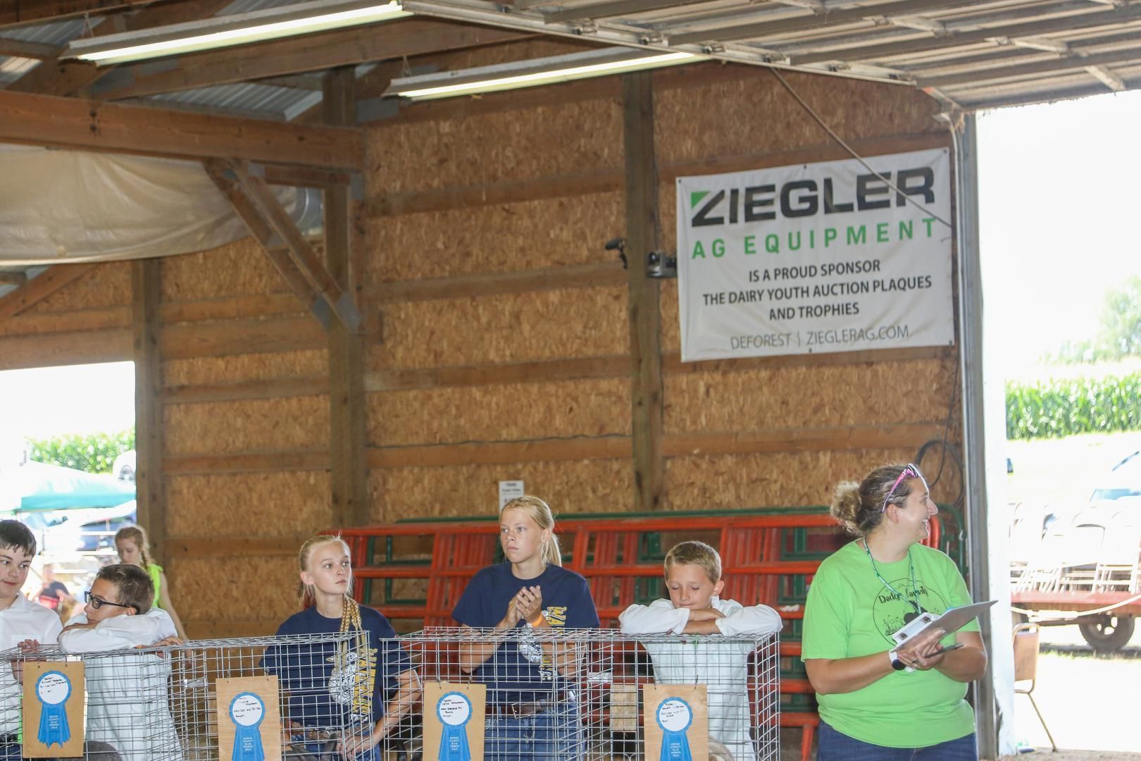 People behind a wire fence at a petting zoo, with a Ziegler Equipment sign on the barn wall.
