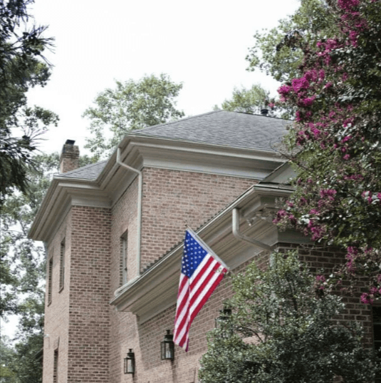 Brick house with an American flag hanging from the front, framed by trees and flowering bushes.