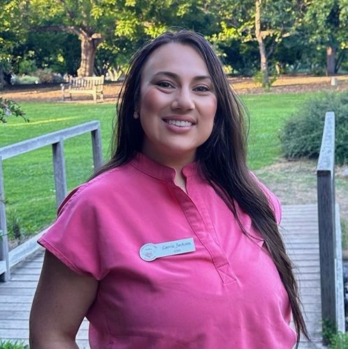 Woman wearing a pink shirt smiles, standing on a wooden bridge, outdoors.