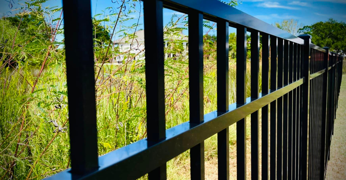 Black metal fence bordering a green grassy area and trees on a sunny day.