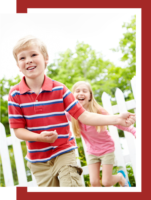 A boy and a girl are playing in front of a white picket fence.