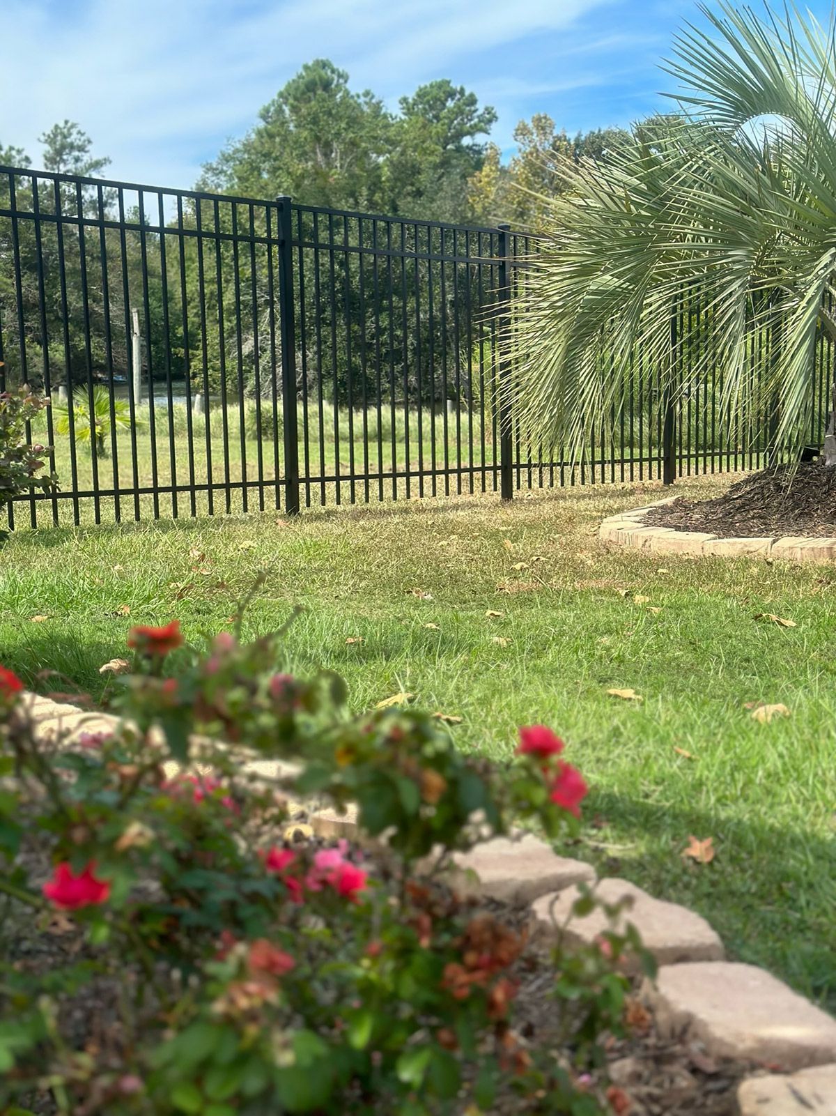 A black fence surrounds a lush green lawn with flowers in the foreground.