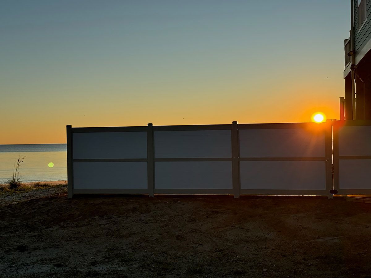 Sunset over a calm body of water, partially obscured by a white fence; a house to the right.