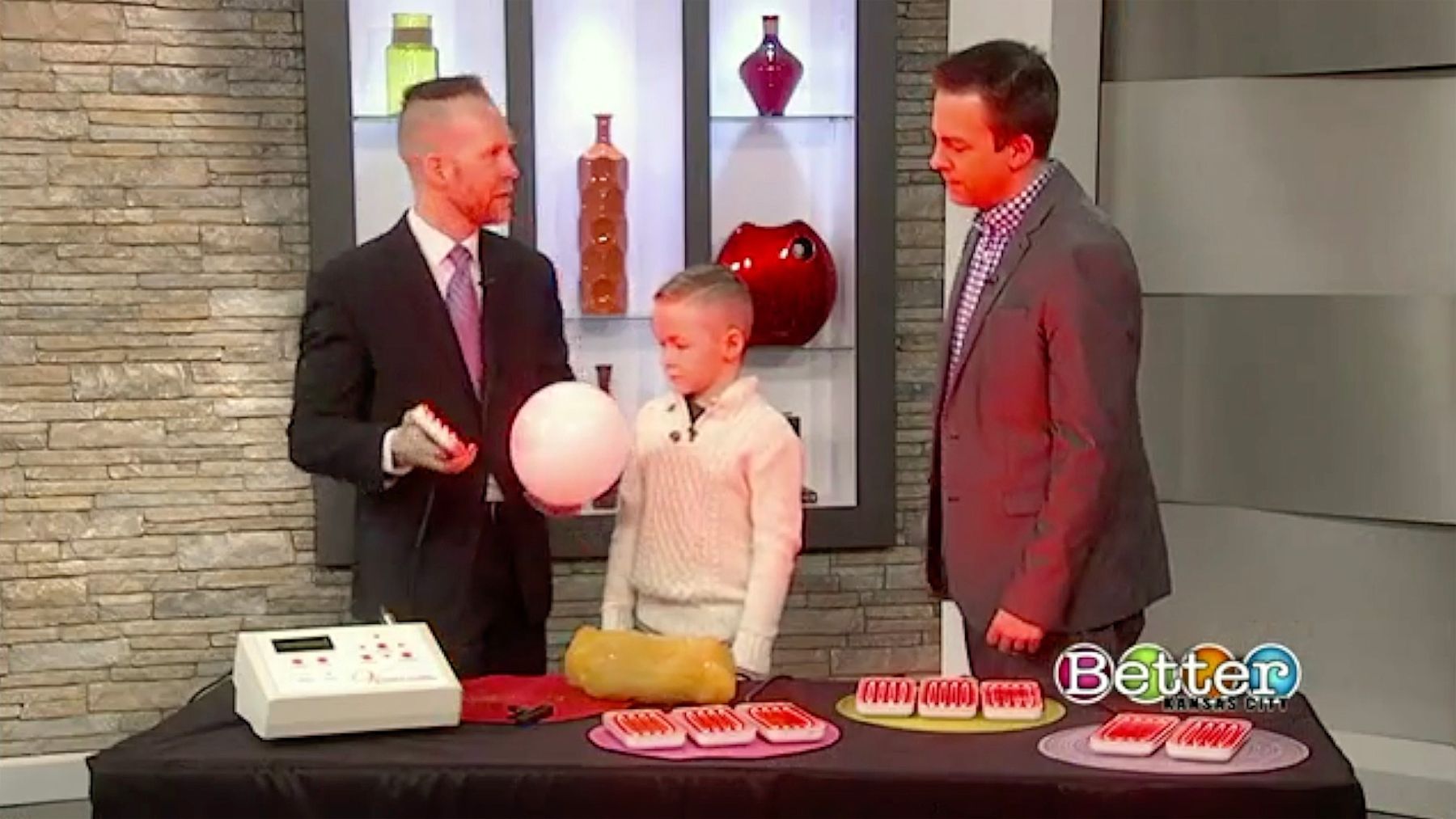 Three men are on a TV set with a boy. The man on the left holds a balloon, gesturing to the child as the other man watches.