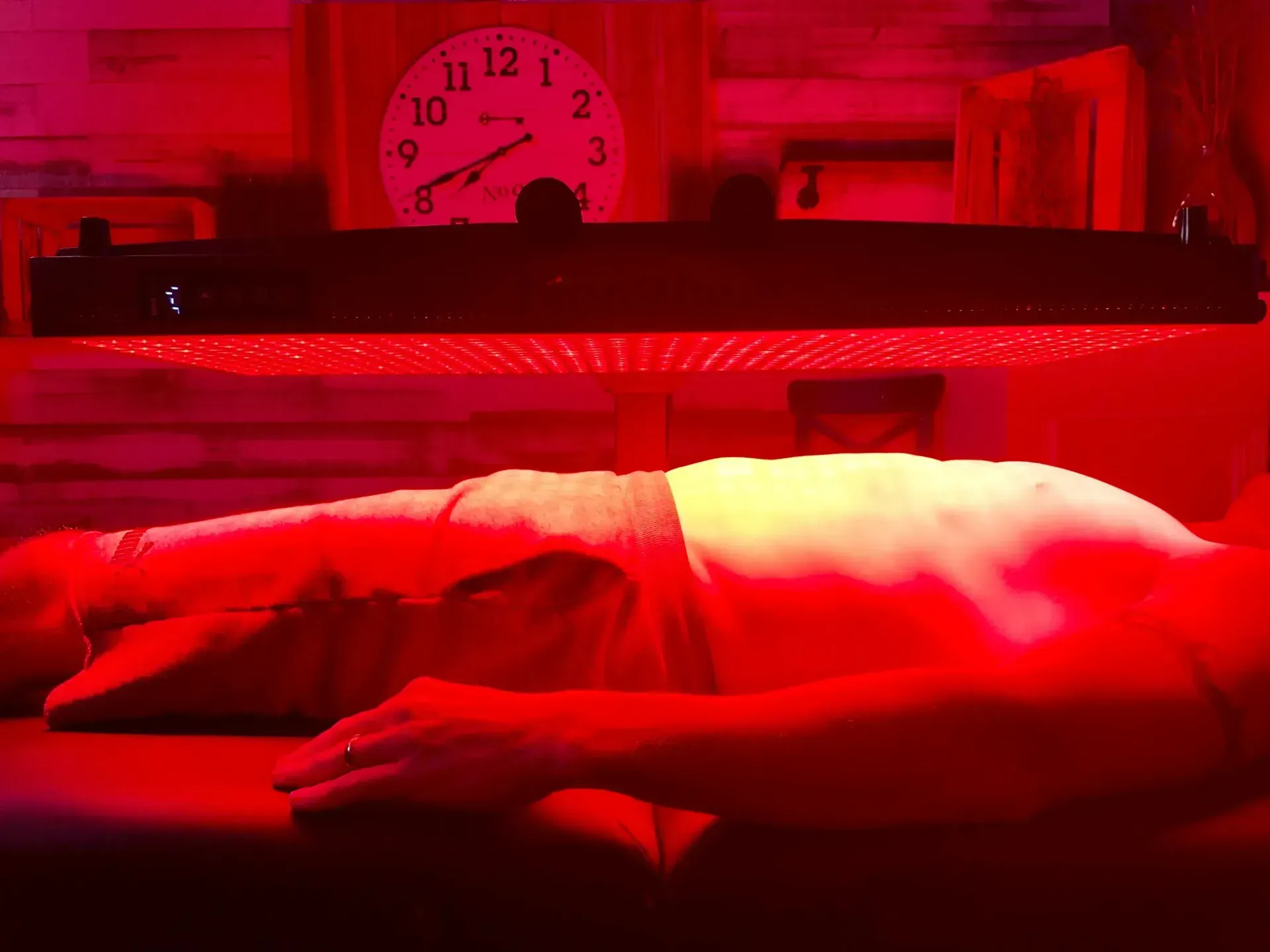 Person receiving red light therapy on their back, lying on a table. Clock on wall.