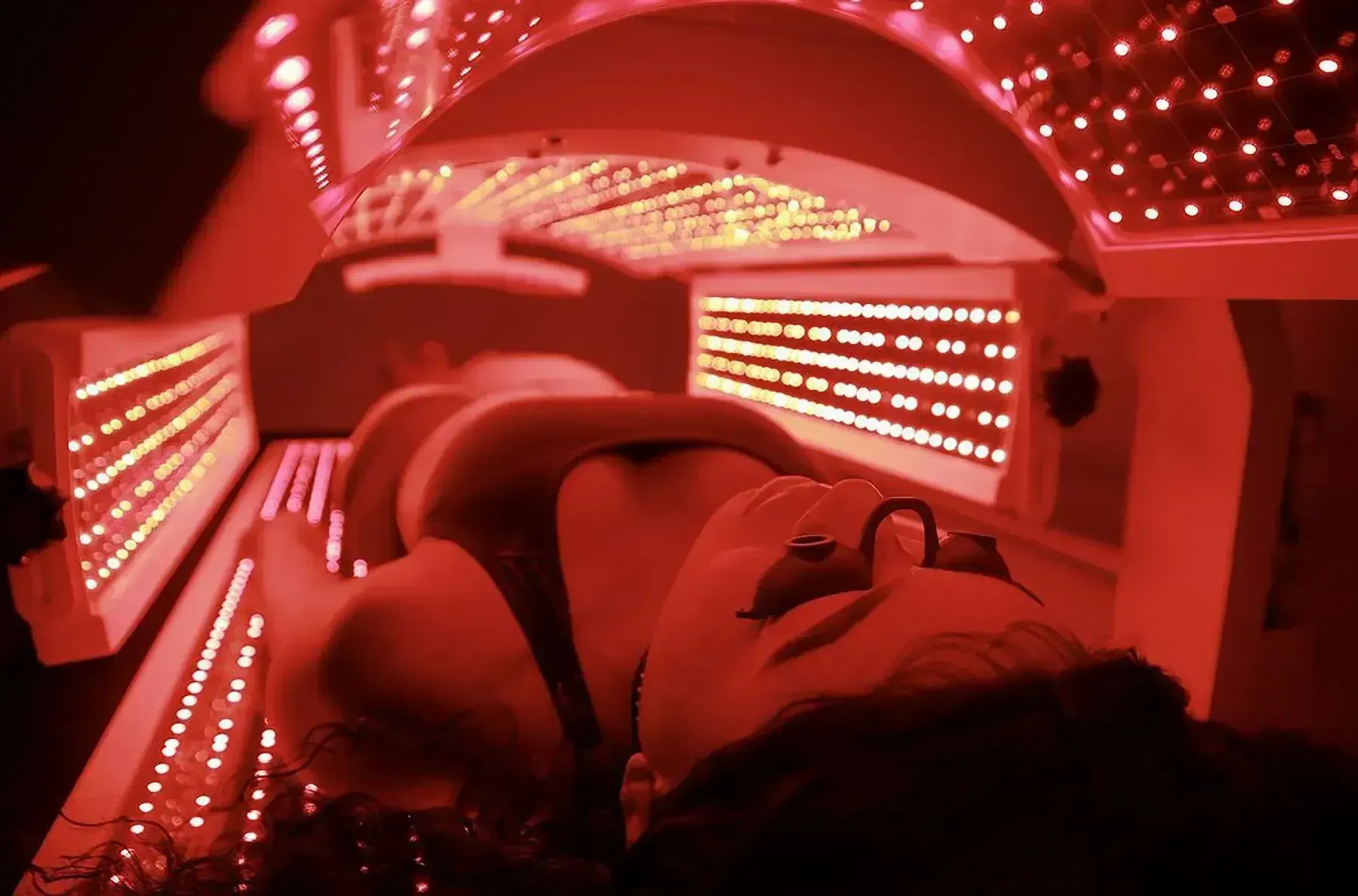 Woman wearing eye protection lies under red LED light therapy machine.