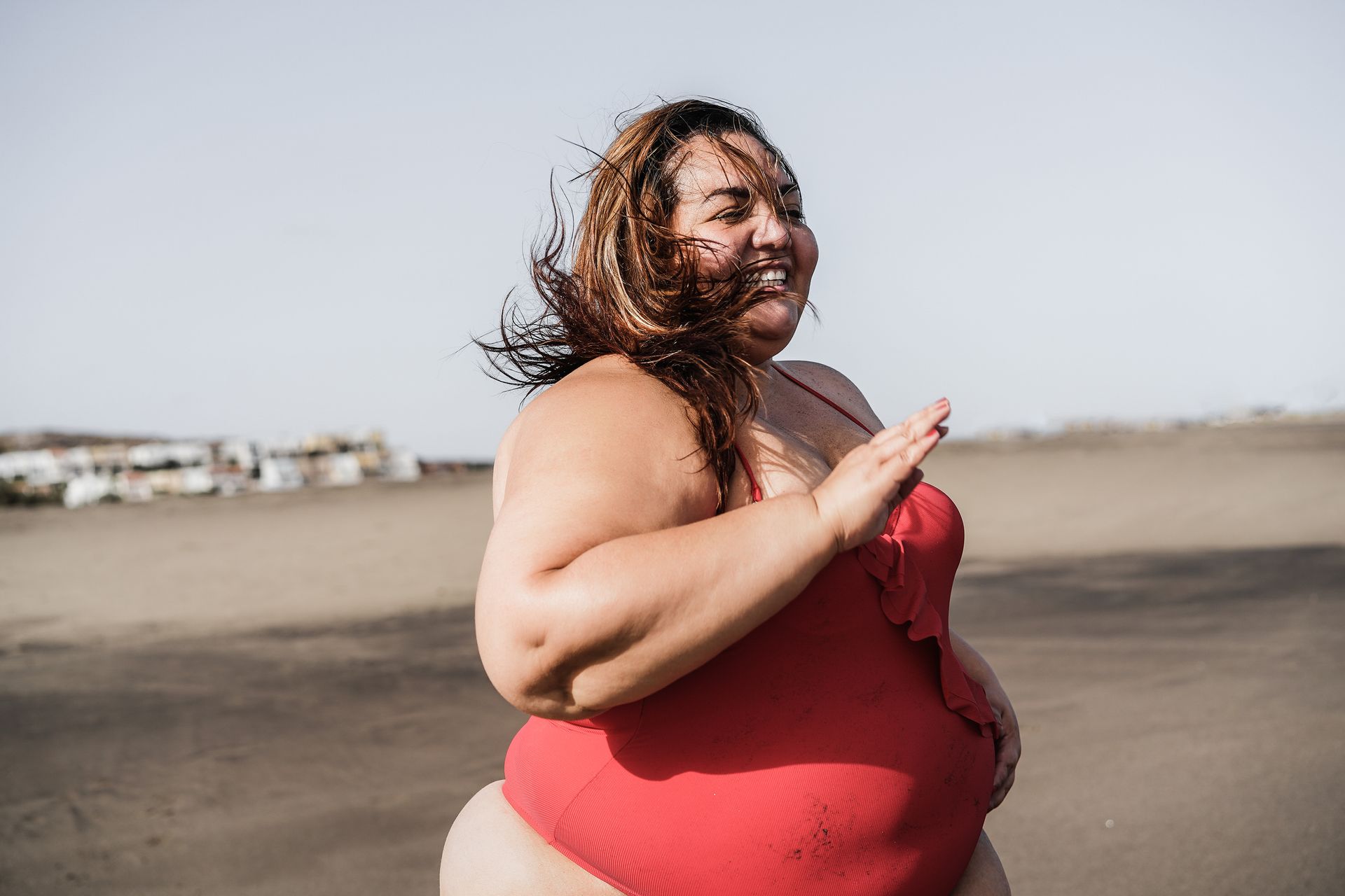 Person in a red swimsuit posing on a beach, smiling in the wind.