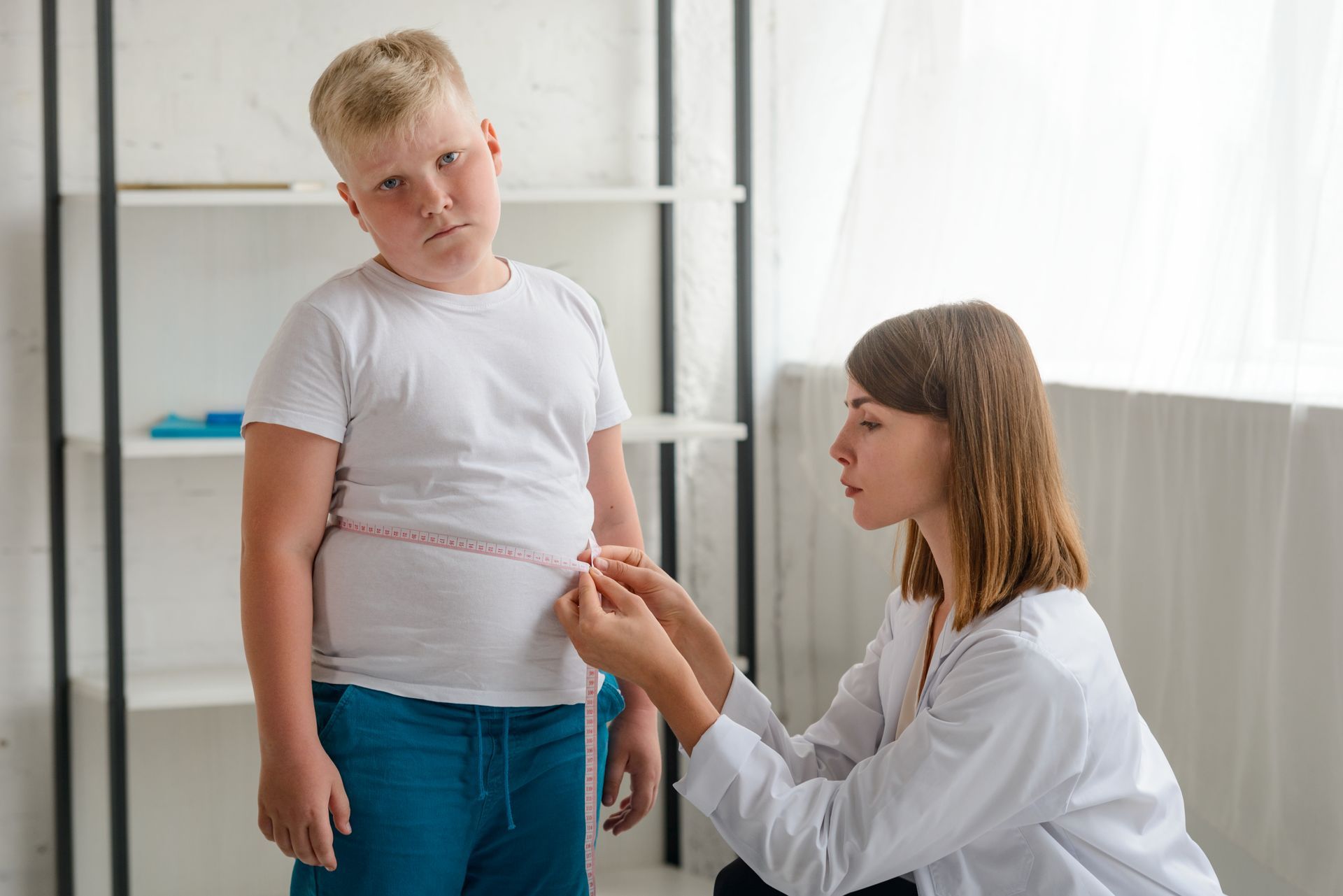 Doctor measuring a boy's waist in a bright room. Boy looks unhappy. White shirt and blue pants.