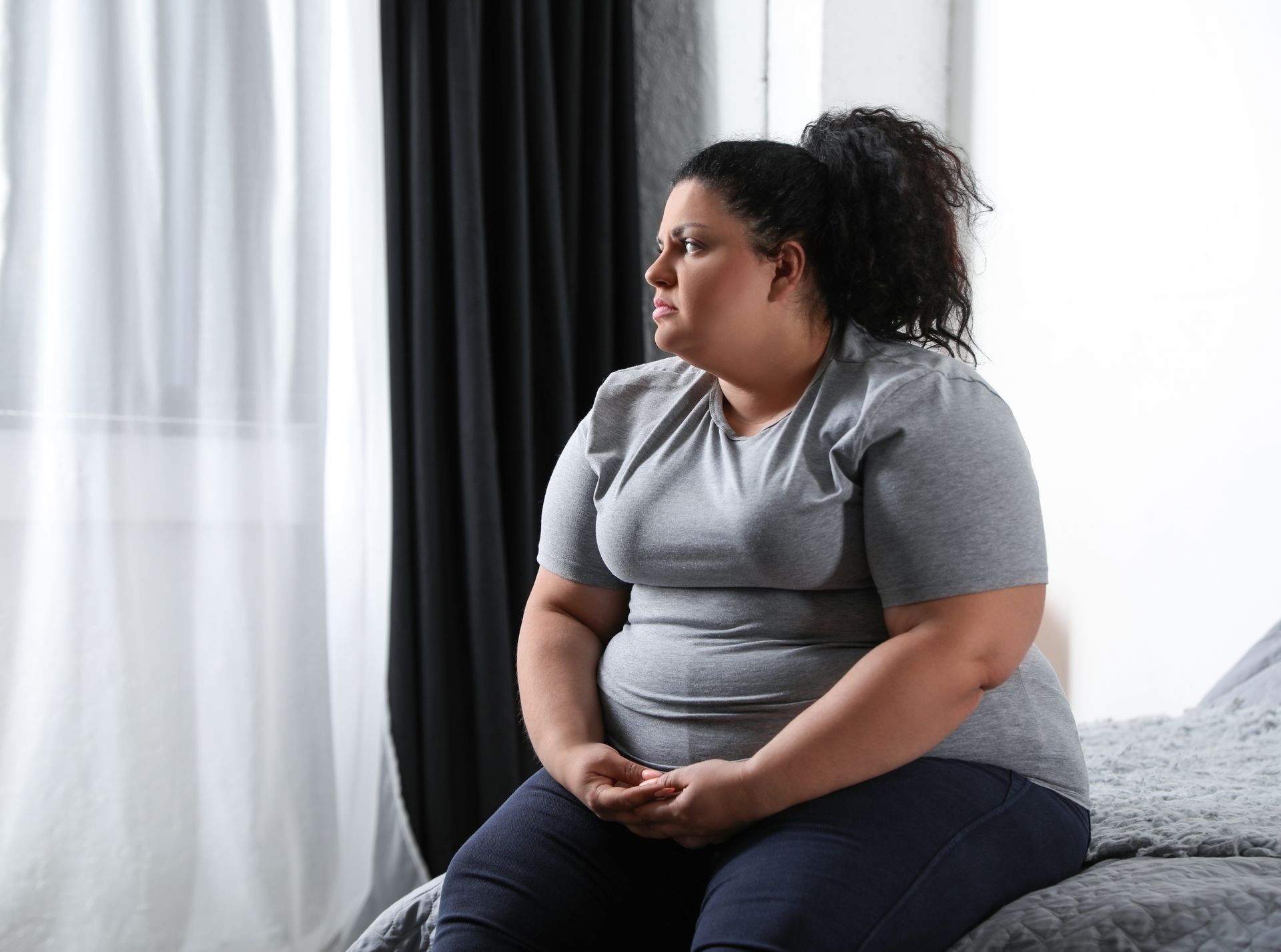Woman sitting on a bed, looking off to the side, wearing a gray shirt and dark pants.