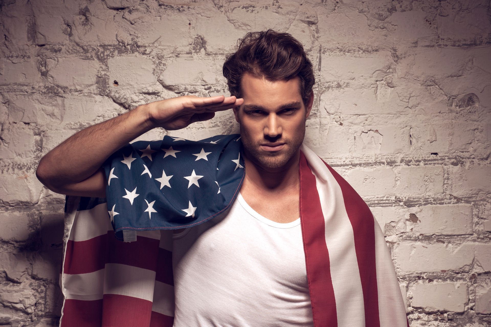 Man saluting, draped in an American flag, standing in front of a brick wall.