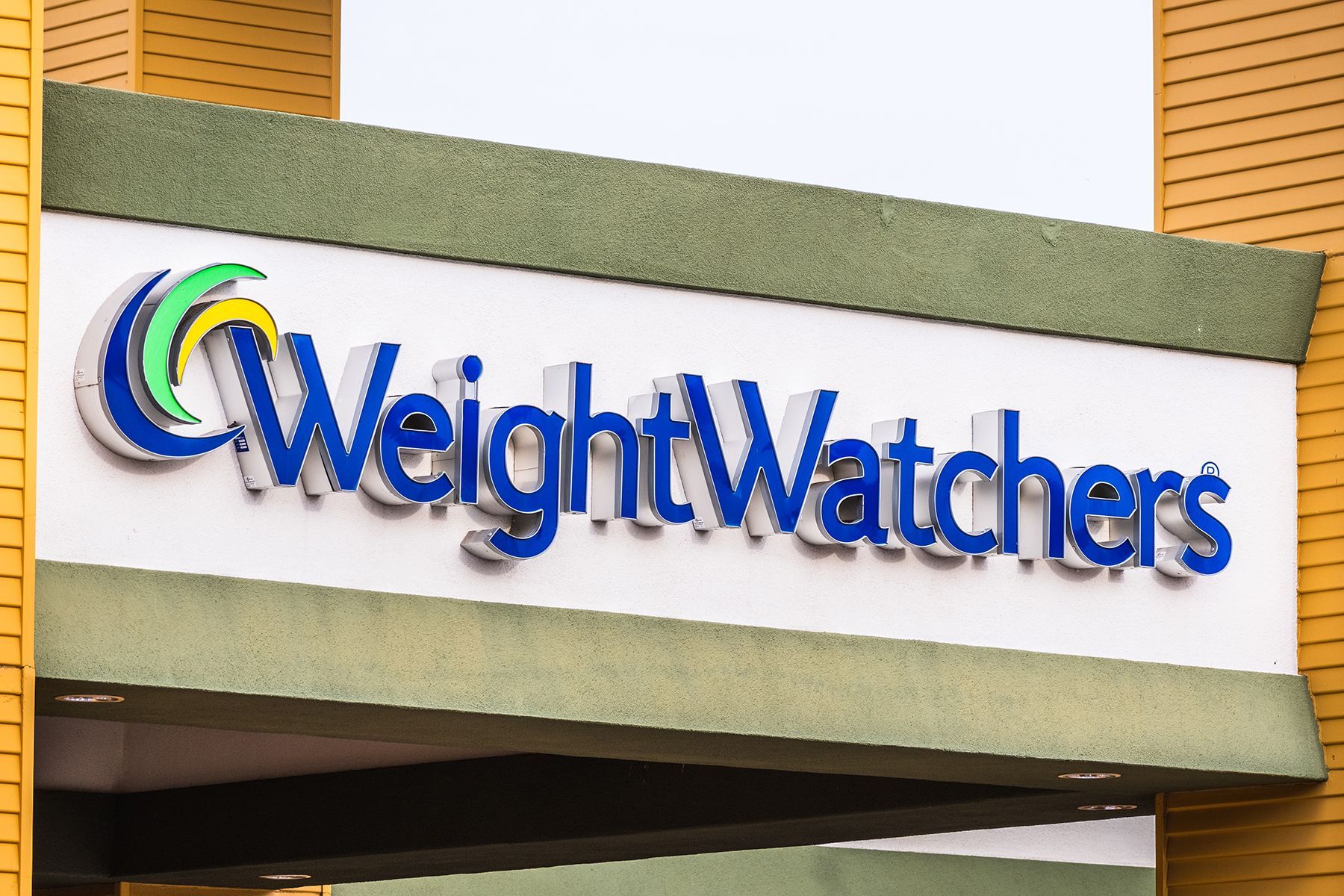 Weight Watchers sign with blue text and logo, on a white and green facade.