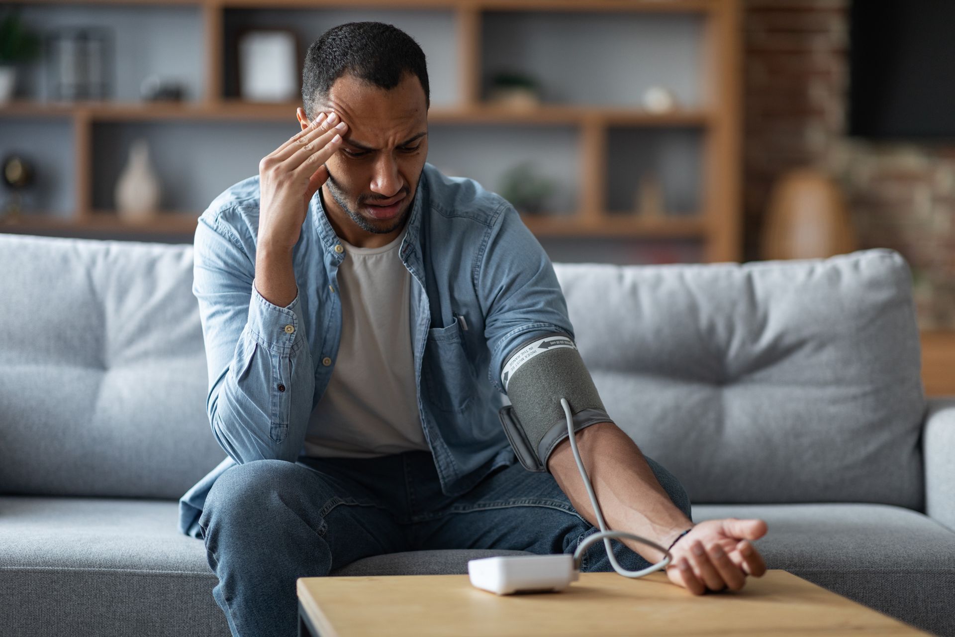 Man on sofa checks blood pressure with a home monitor, holding his head in discomfort.