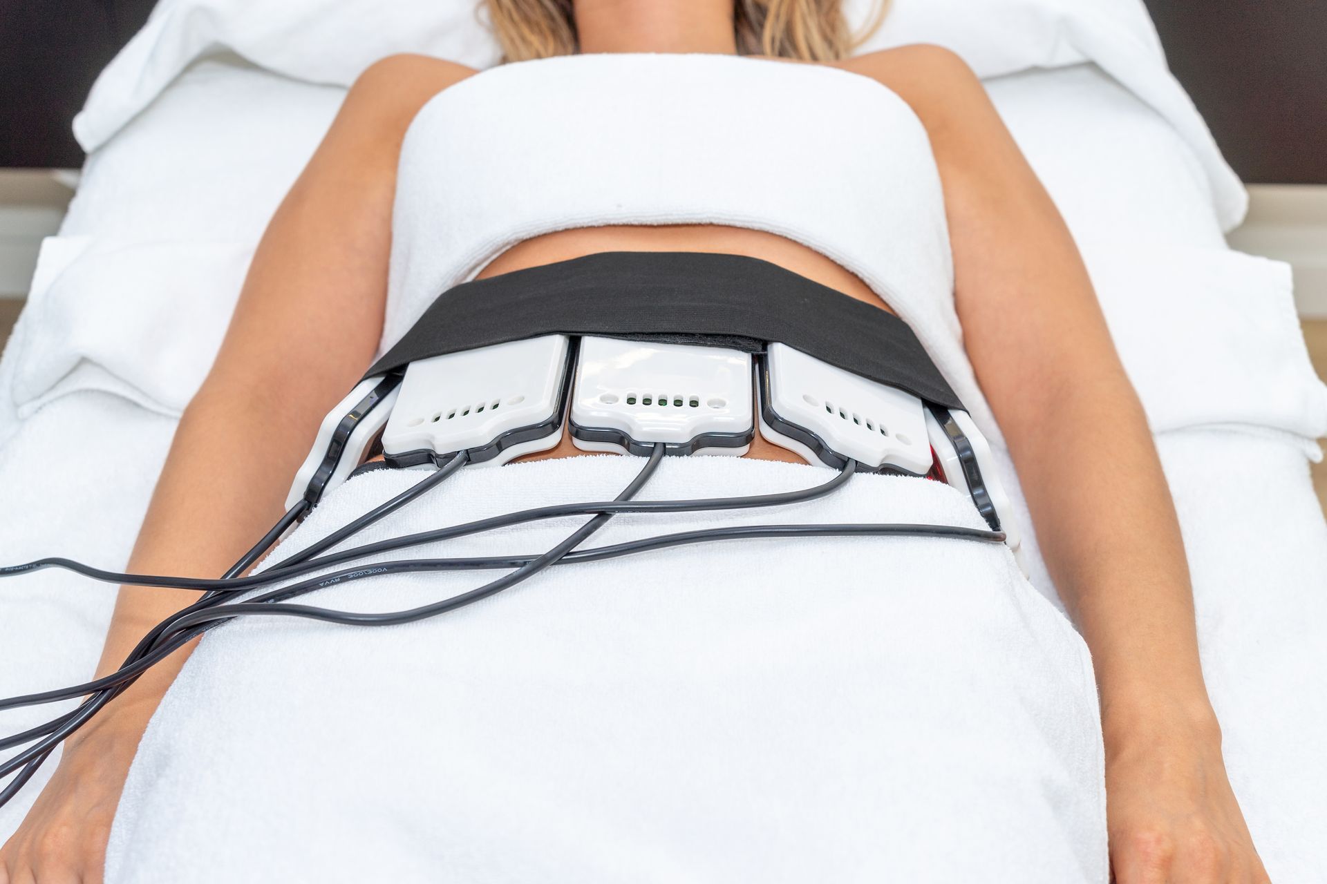 Woman lying down with a black belt and connected devices around her midsection, undergoing a treatment.