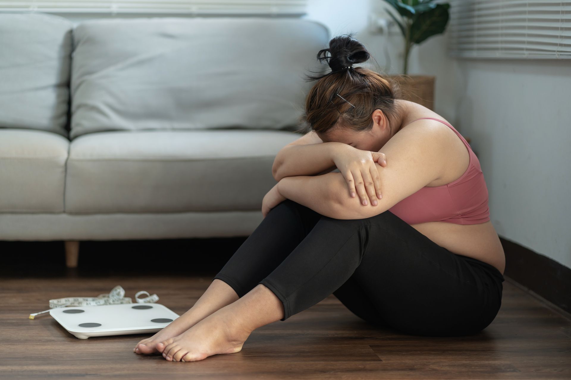 Woman sits on floor, hugging knees, near scale; defeated expression.