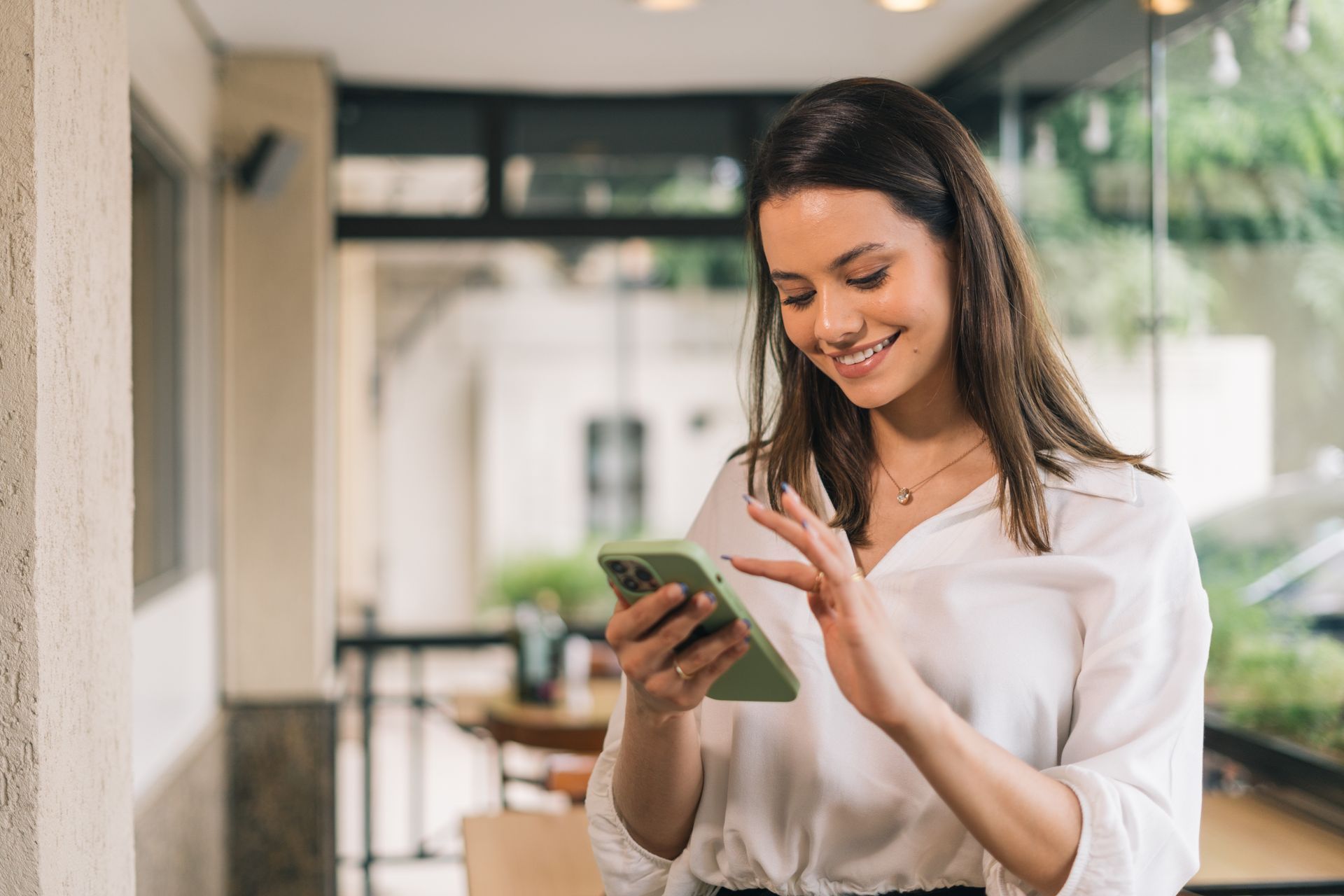 Woman in white shirt smiles while using her phone indoors, near a window.