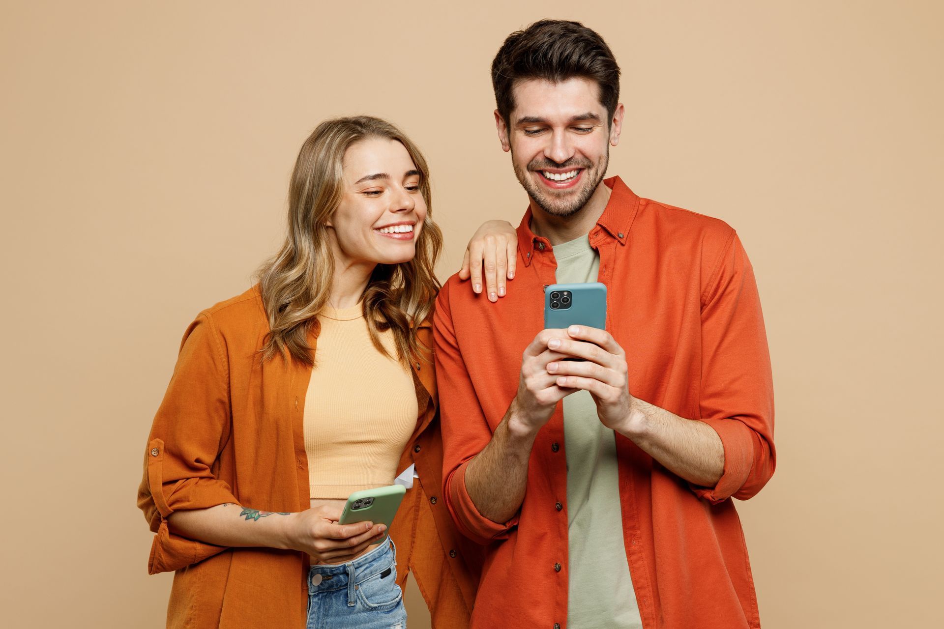 Woman and man smiling, looking at phones. The man holds a phone, the woman's arm rests on his shoulder.