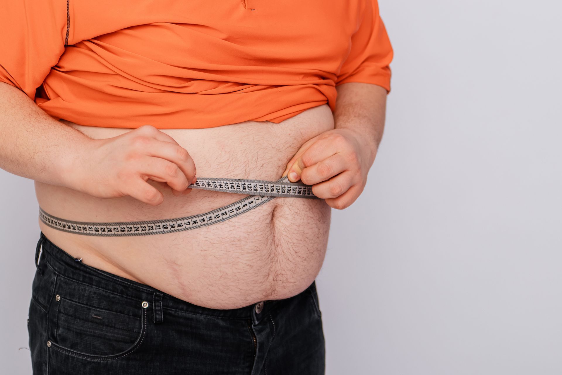 Person measures their waist with a tape measure, belly visible; orange shirt, dark pants.