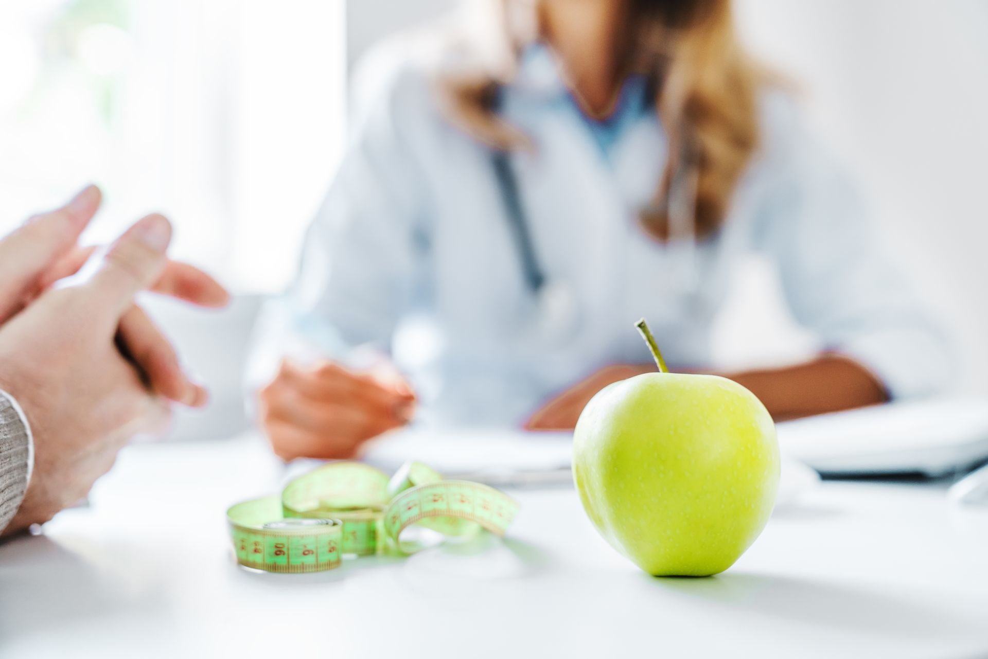 Green apple, measuring tape, and blurred doctor consulting with a patient at a desk.